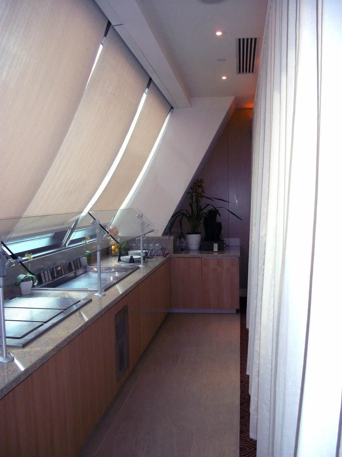 A modern hotel buffet area with a granite countertop, serving trays with covers, and a potted plant at the end of the counter. Beige curtains hang beside the buffet area, with ceiling lights and an air vent visible.