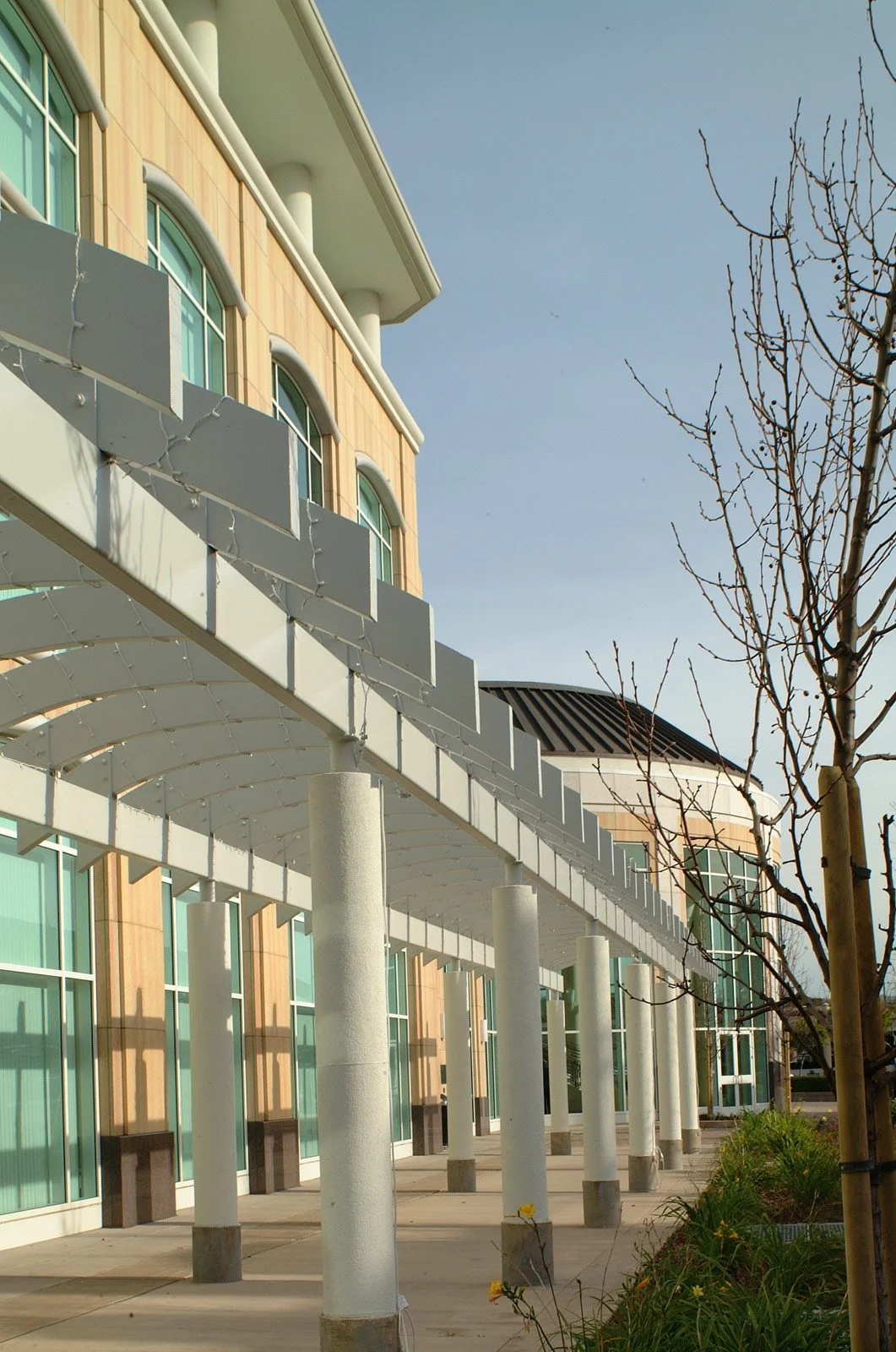 Exterior of a modern building with large windows, white columns, and a walkway with supporting pillars, under a clear blue sky.