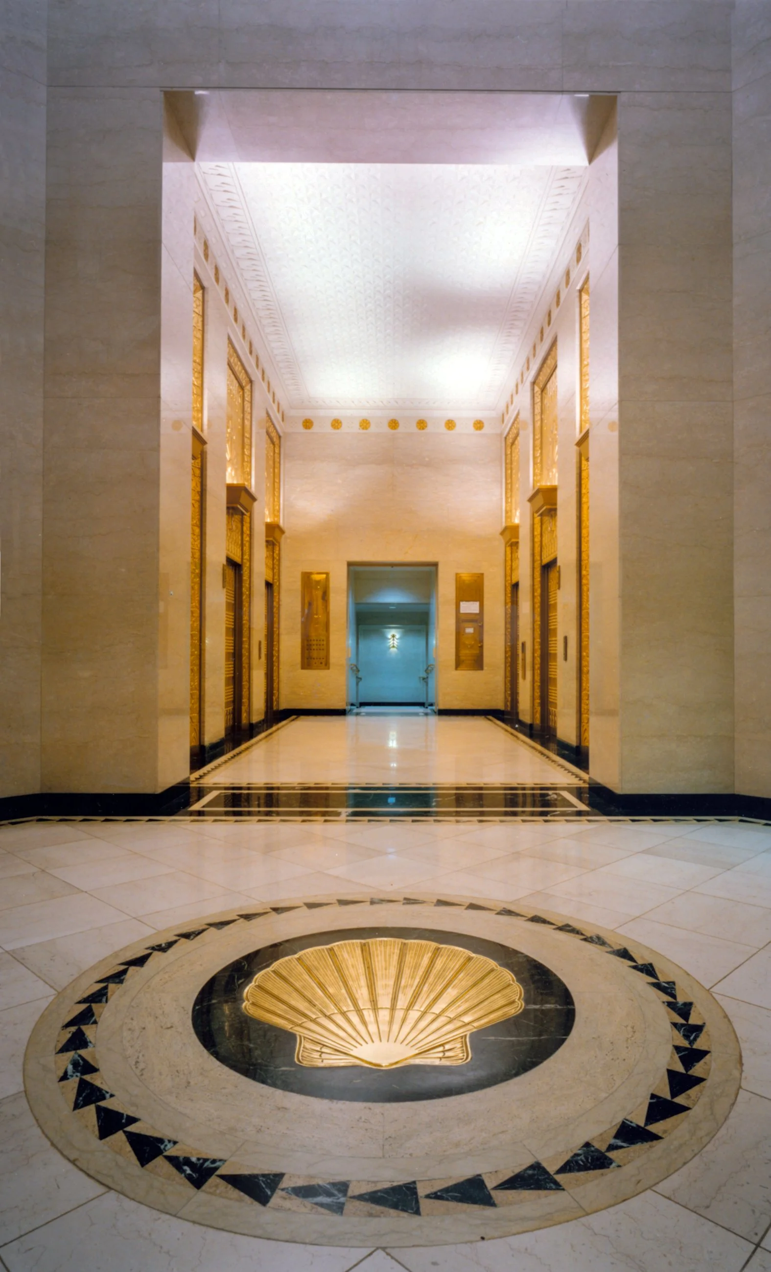 Elegant hotel lobby with marble flooring, golden accents, and elevator doors, featuring a decorative shell design on the floor in the foreground.