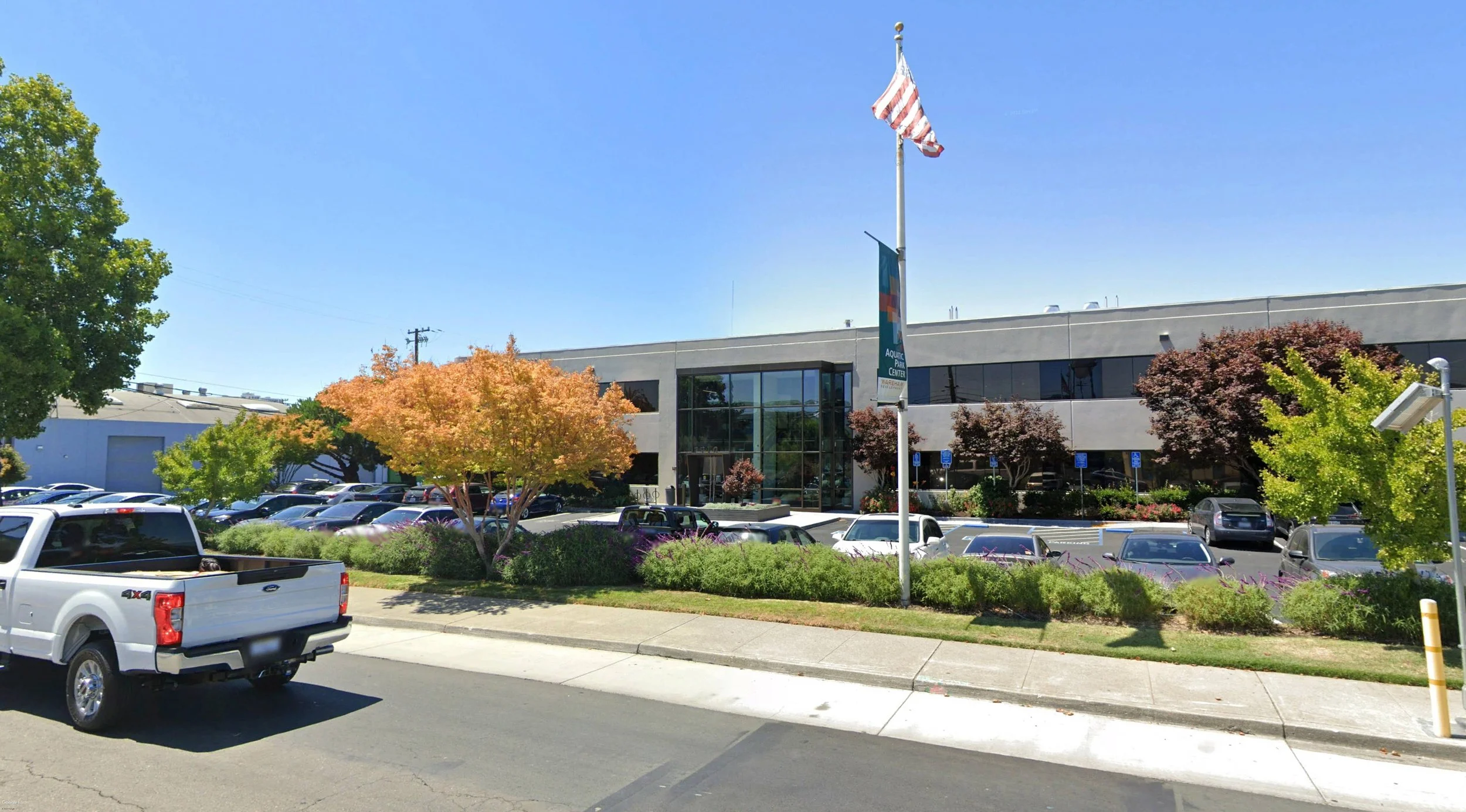 Parking lot with cars in front of a commercial building, trees, and blue sky.