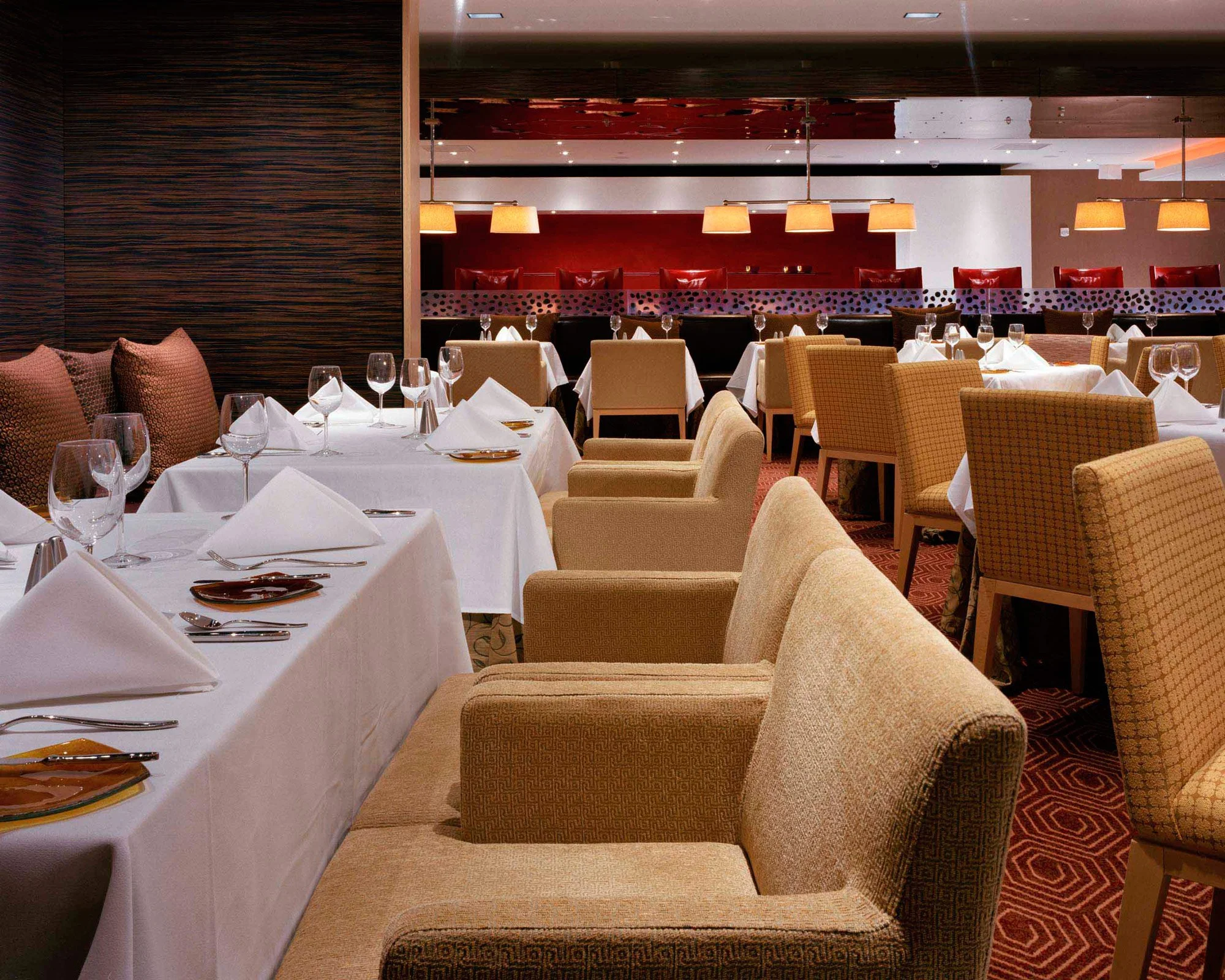 Elegant restaurant dining room with tables set with white tablecloths, napkins, wine glasses, and plates, surrounded by beige and brown upholstered chairs, with warm lighting and red accent wall.