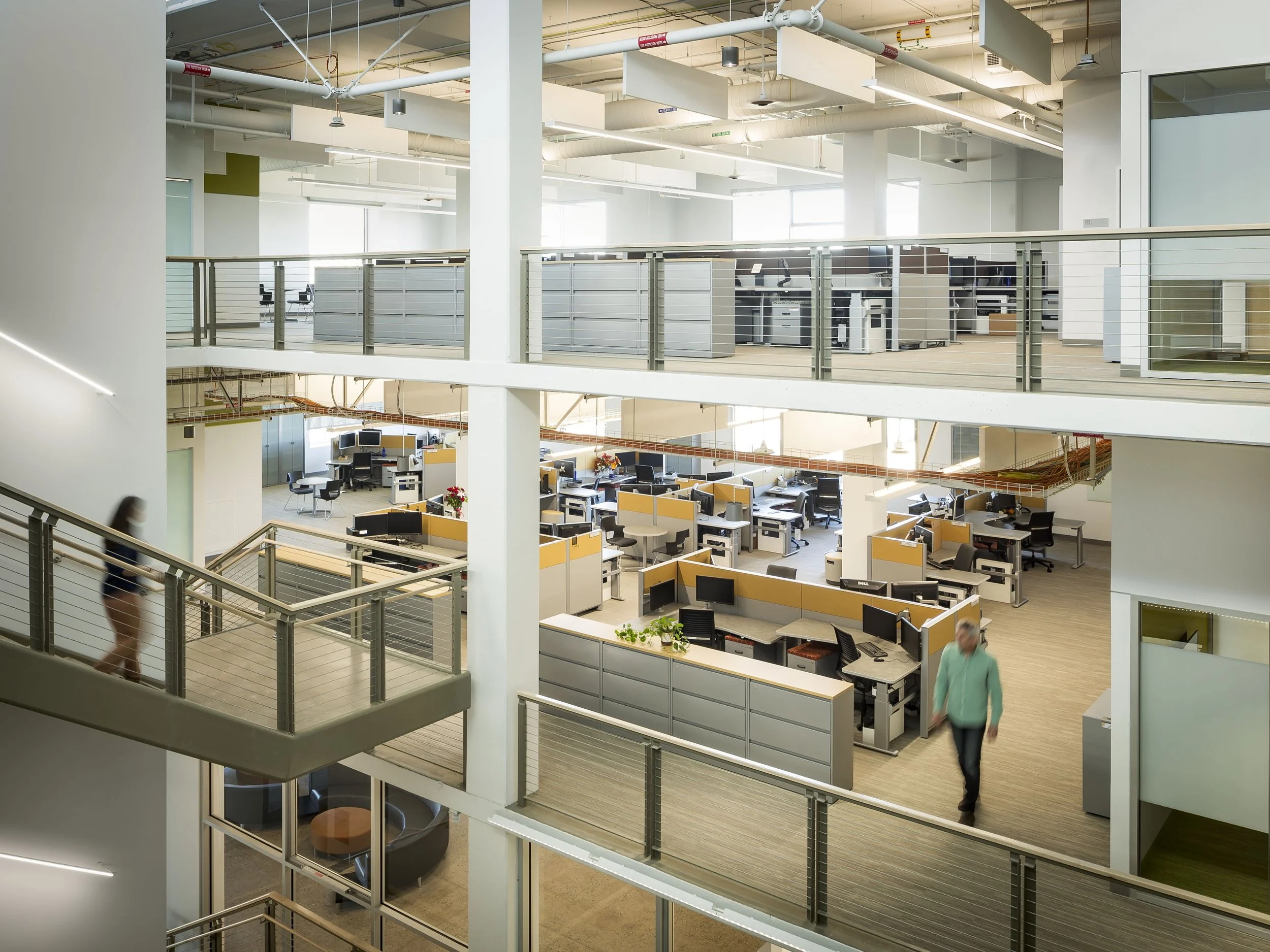 Open-plan modern office with multiple workstations, partition cubes, and two levels connected by walkways, with people walking and working.
