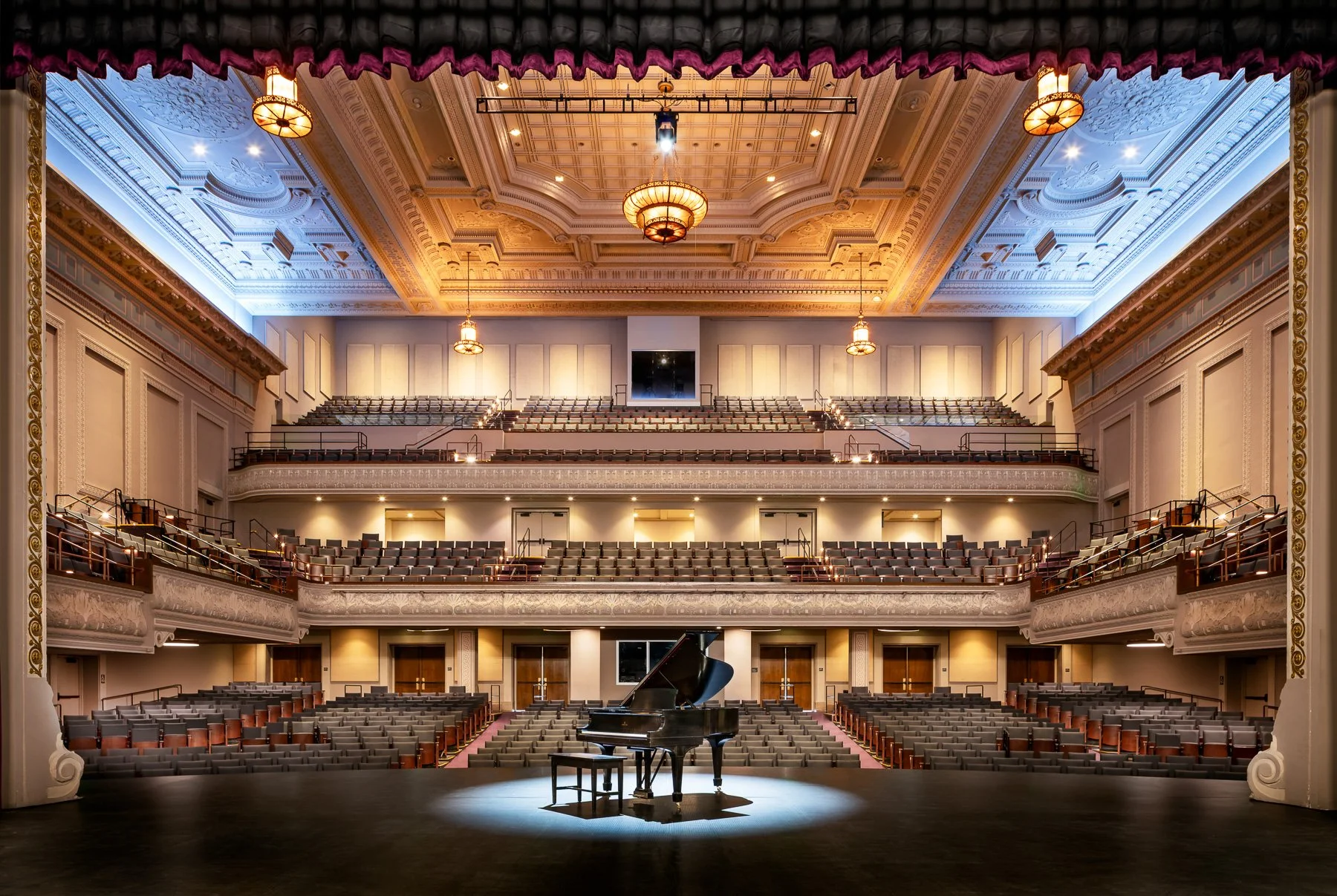 Interior view of an ornate concert hall with a grand piano on stage, surrounded by multiple tiers of empty seating and elegant chandeliers hanging from the decorated ceiling.