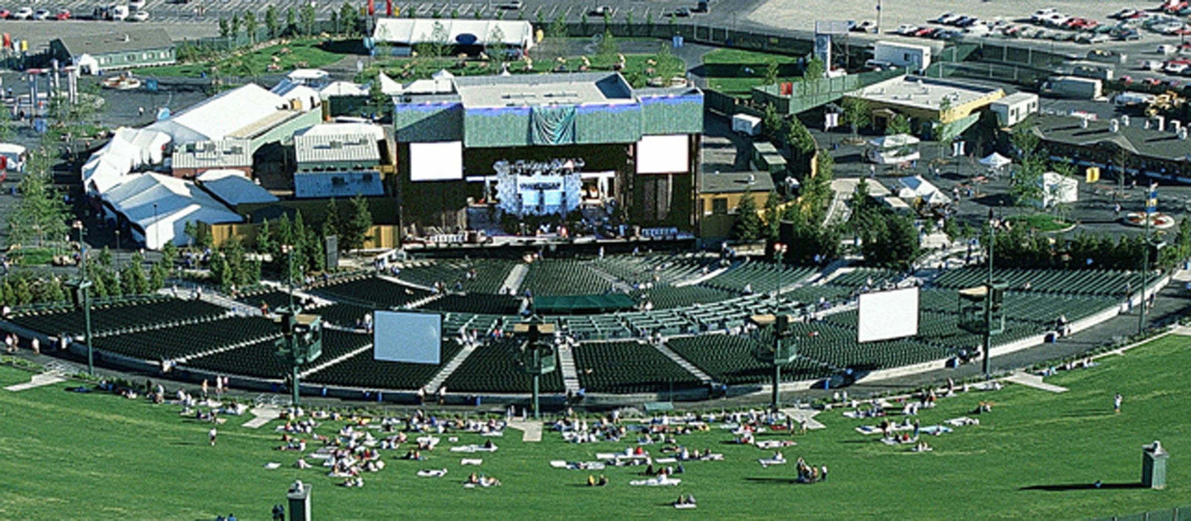 An outdoor concert venue with a stage and seating area surrounded by a lawn where many people are sitting or lying down on blankets. Tents and parking lots are visible in the background.