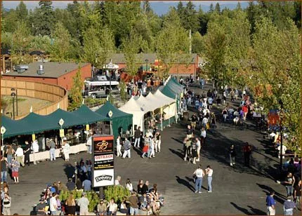 A busy outdoor market or fair with multiple tents, vendors, and people walking around, set against a backdrop of trees and buildings.