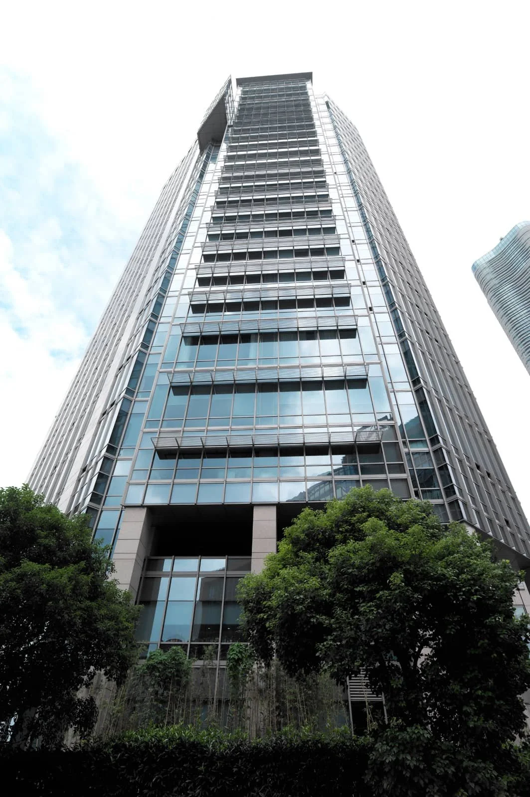 Tall modern glass office building with trees at the base, reaching up toward a cloudy sky.