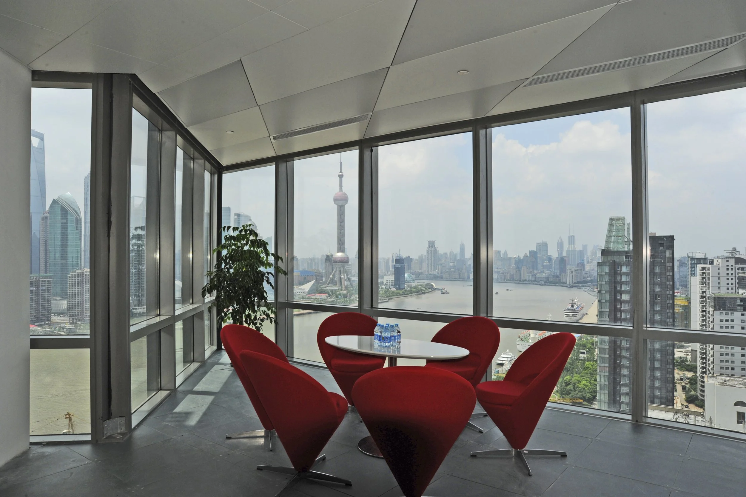 Indoor meeting area with five red chairs around a white round table, with bottled water, in a high-rise with large windows showing a city skyline, including the Oriental Pearl Tower and a river.