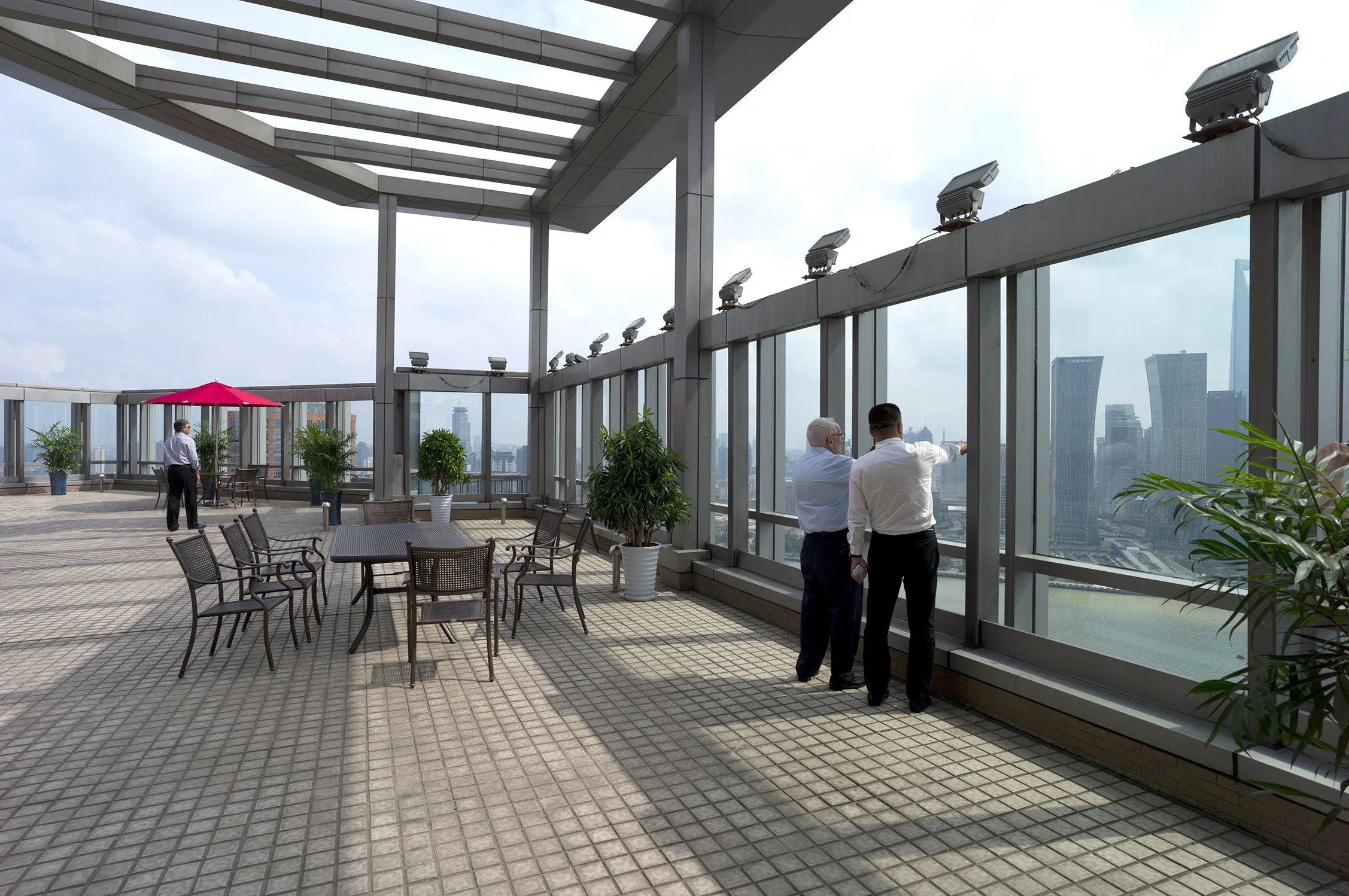 Businesspeople on an outdoor rooftop terrace with city skyline view, glass walls, potted plants, tables, chairs, and a red umbrella.