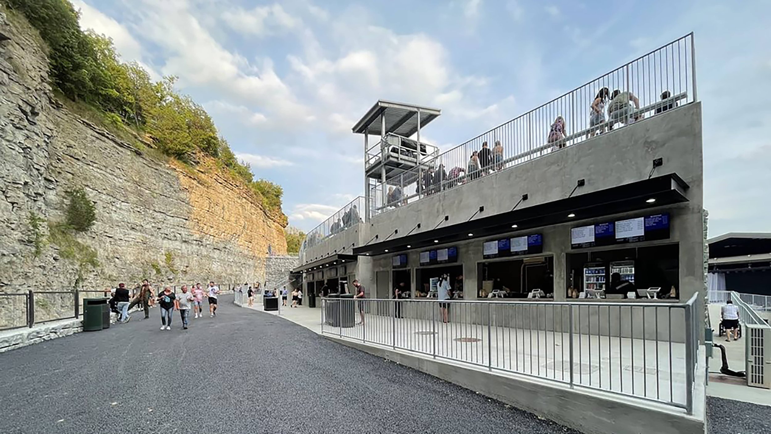 People at a waterfront outdoor venue with a modern concrete building, multiple digital screens, and a viewing platform on top, adjacent to a cliff with trees.