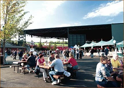 People sitting at picnic tables at an outdoor event, with a stage and tents in the background on a sunny day.
