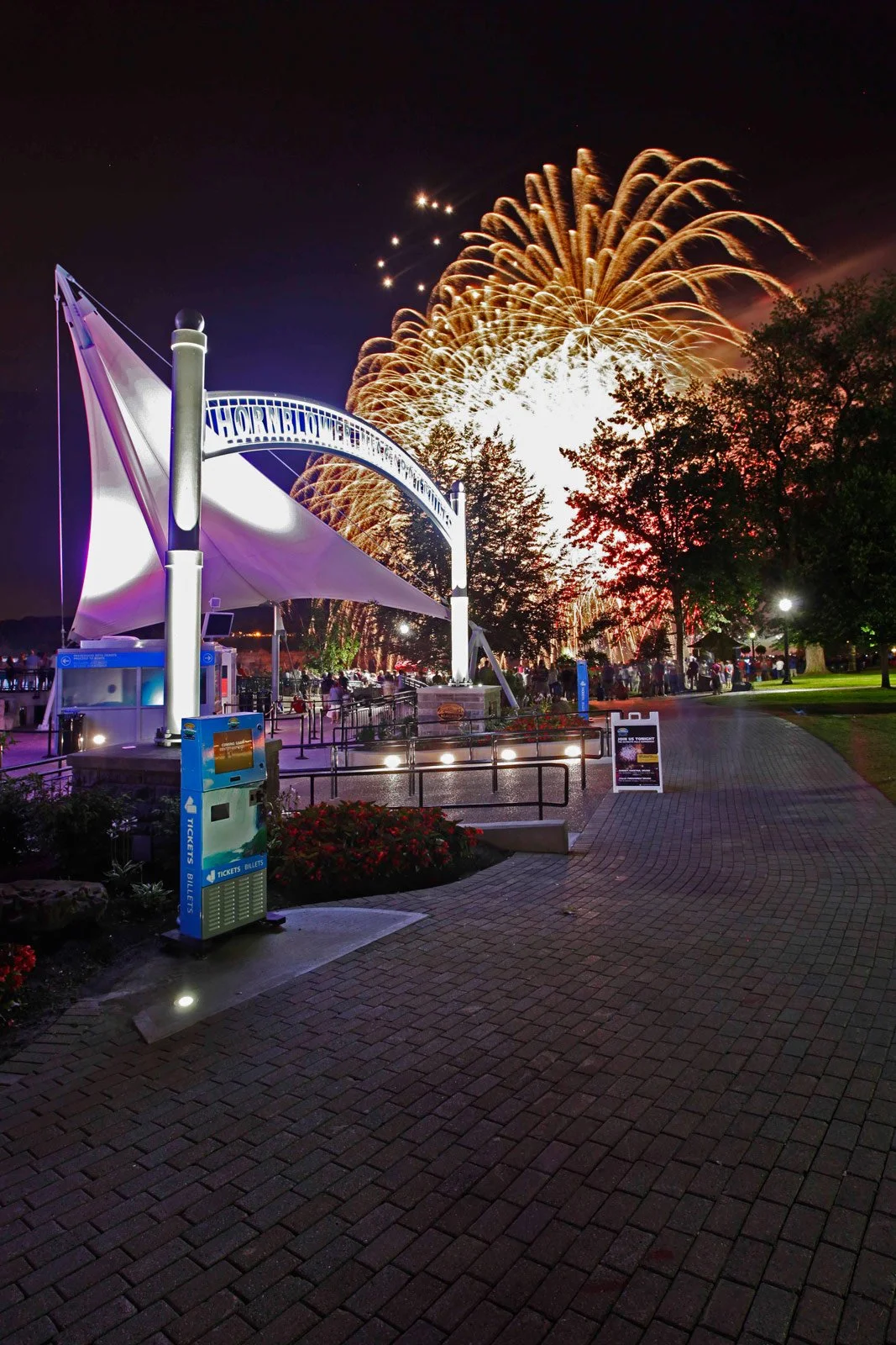 Fireworks light up the night sky over an amusement park with a large sign that reads 'Hornblower' and a canopy-covered ticket booth in the foreground, with several people walking around.