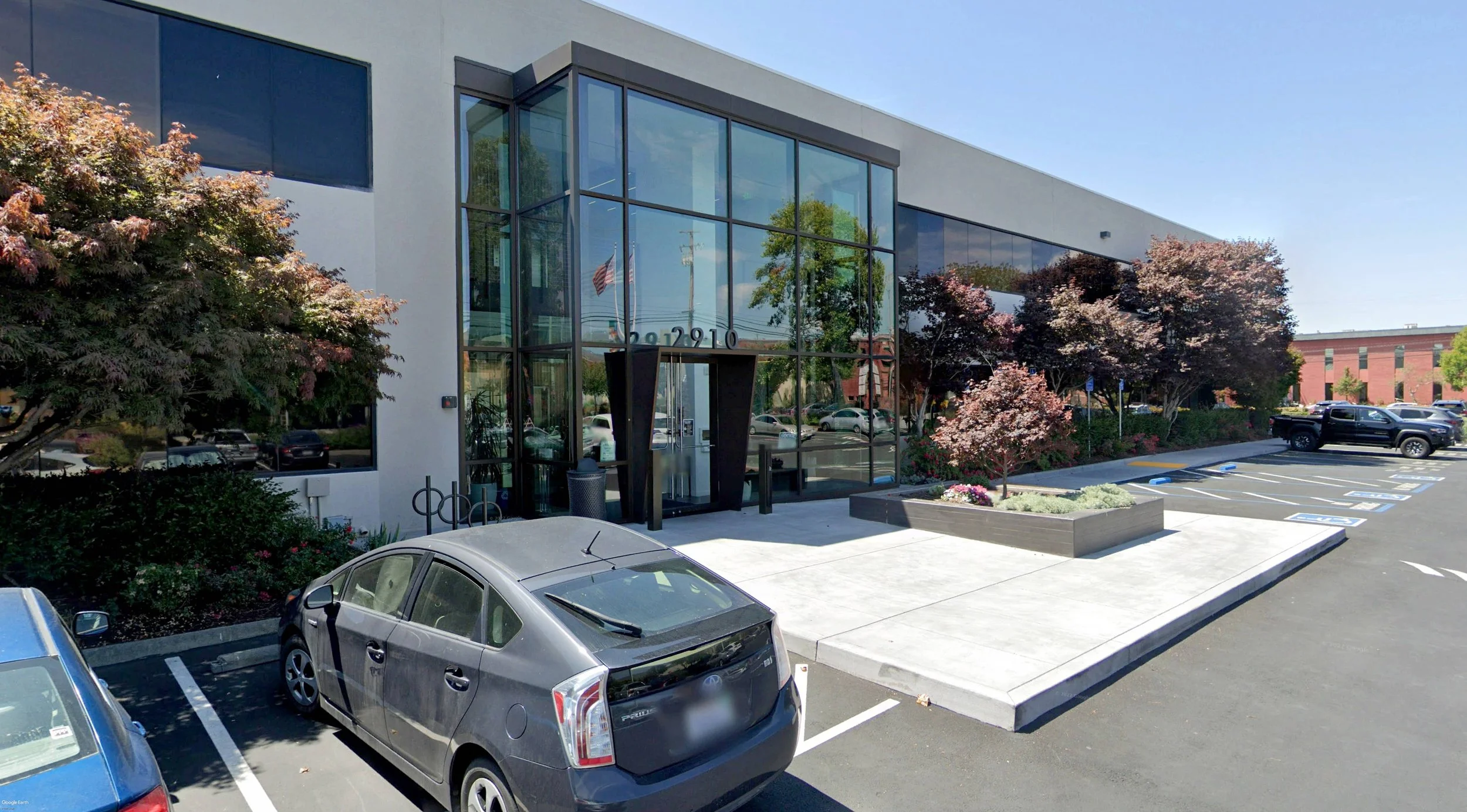 Modern office building with large glass windows and a glass entrance, surrounded by landscaped trees and parking lot with several cars.