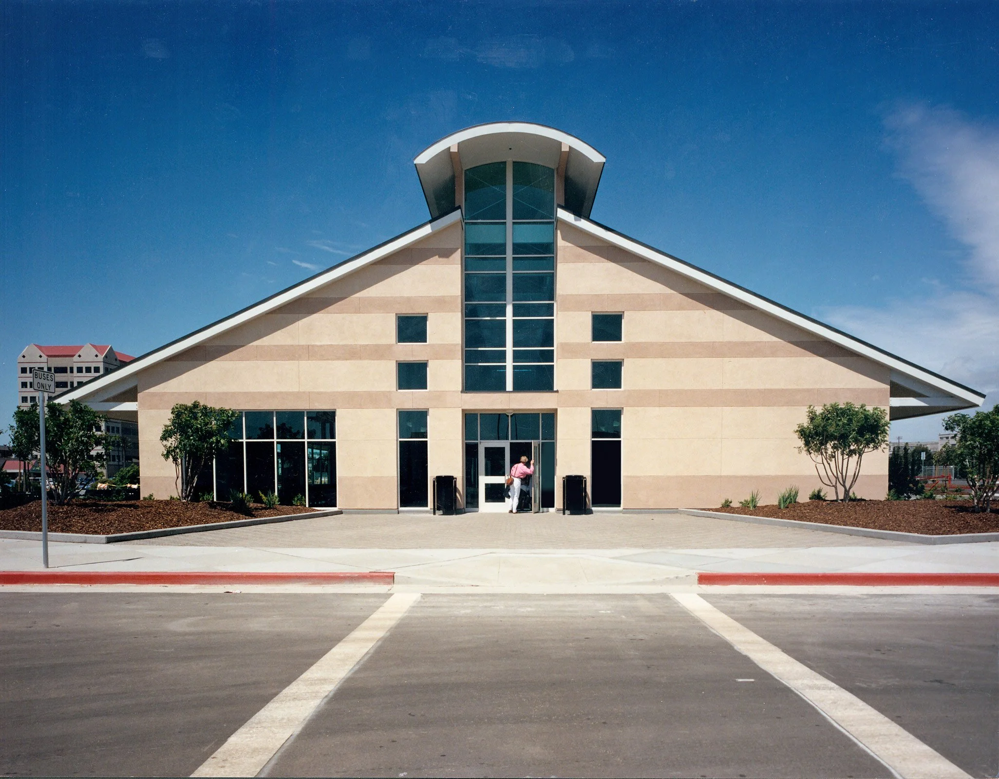 Modern church building with a sloped roof, large central glass window, and beige exterior walls. Two small trees are outside, and a person is entering the door. The parking lot is in the foreground with clear, marked spaces.