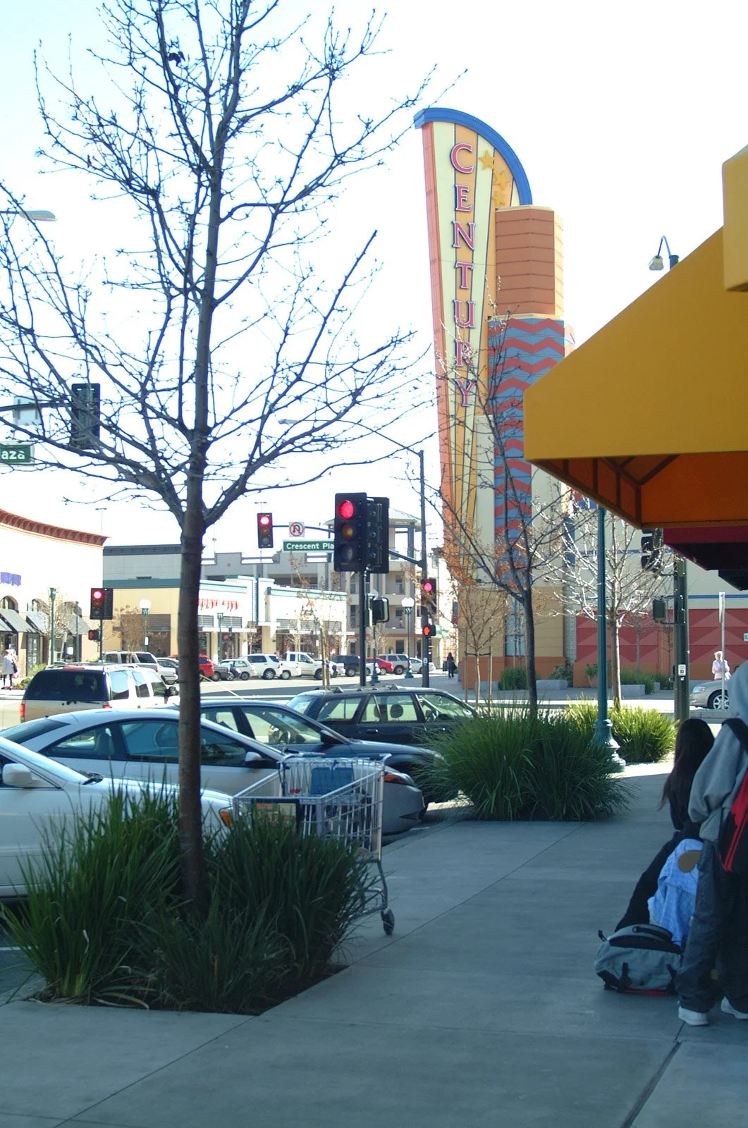 View of a street scene outside of an entertainment or shopping center, with a sign reading 'CENTURY' on a tall building, parked cars, traffic signals, and pedestrians on the sidewalk.