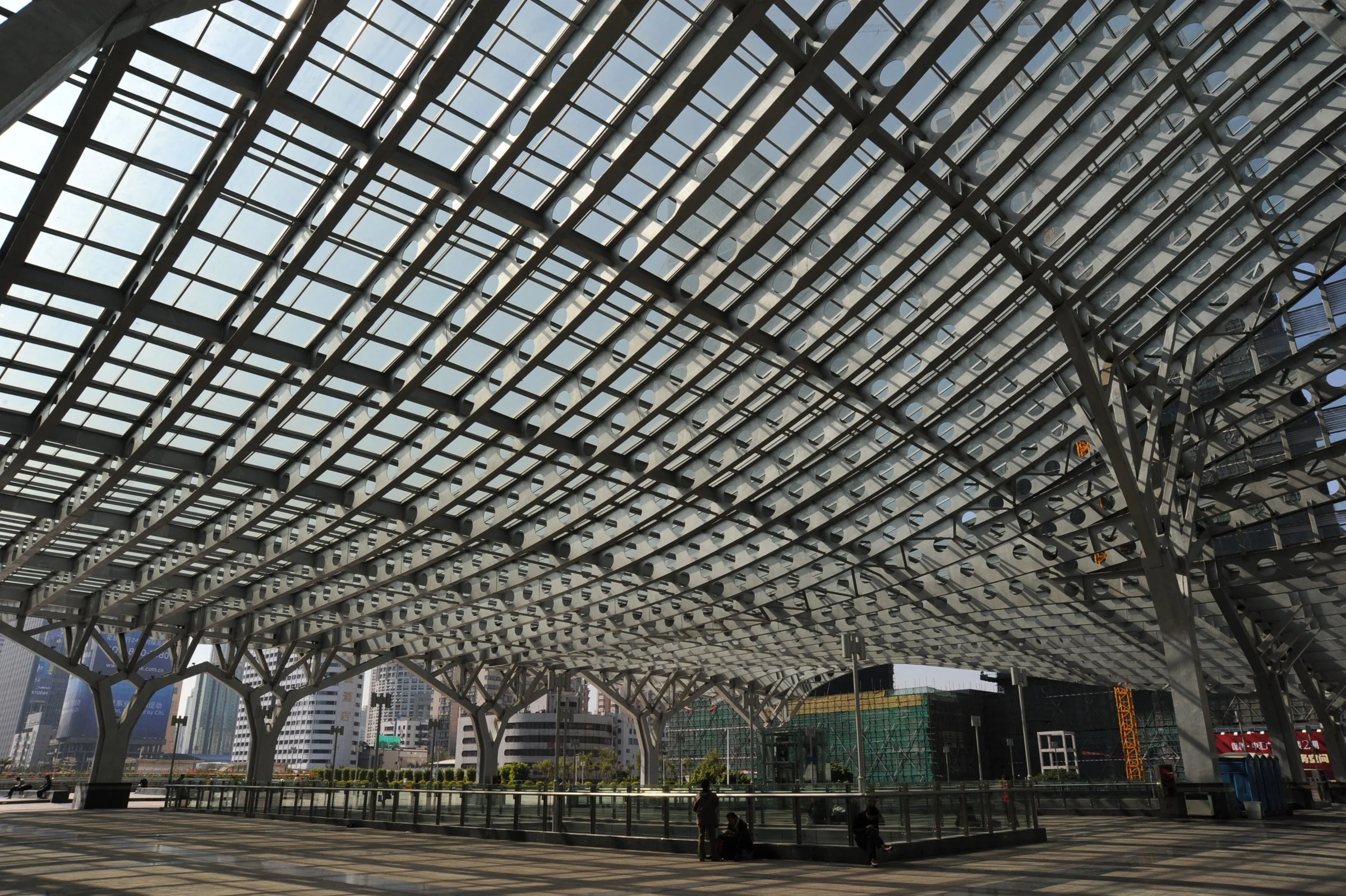 Modern architectural structure with a complex metal and glass roof, beneath a city skyline with tall buildings.