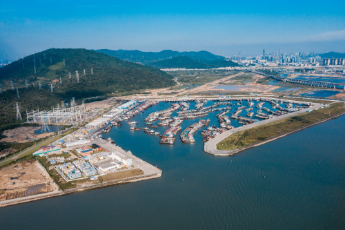 Aerial view of a marina with boats docked, surrounded by hilly terrain and urban area in the distance.