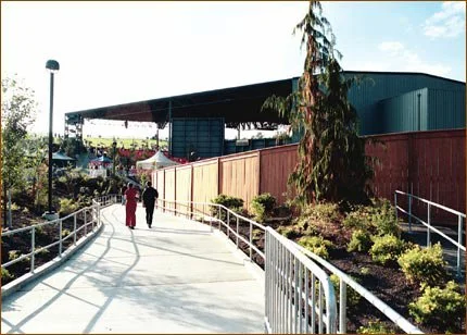 People walking on a paved outdoor path with railings, nearby plants, a building with a blue exterior, and a clear sky.