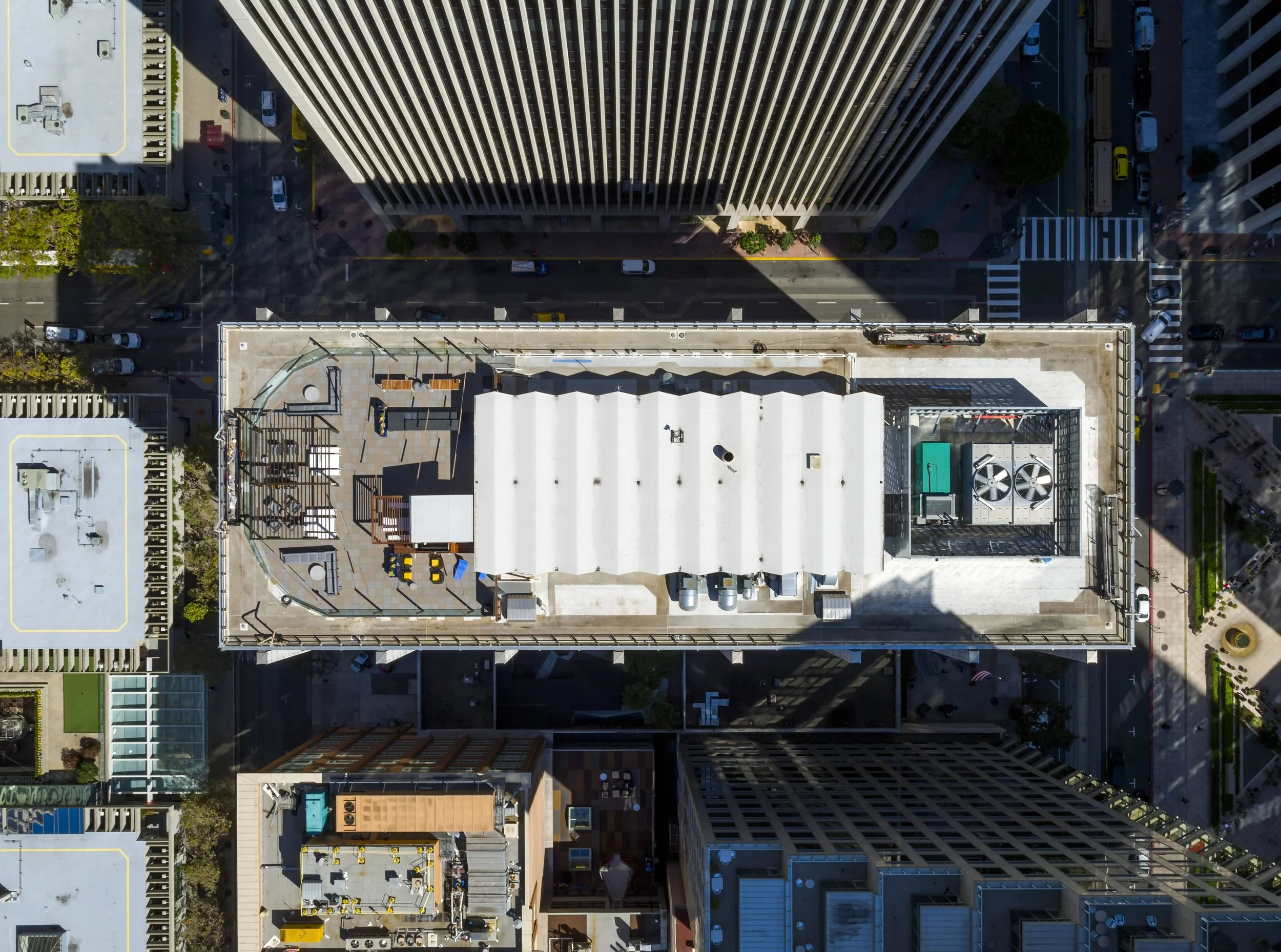 Aerial view of city rooftops with tall skyscrapers, streets, vehicles, and parking areas below on a sunny day.