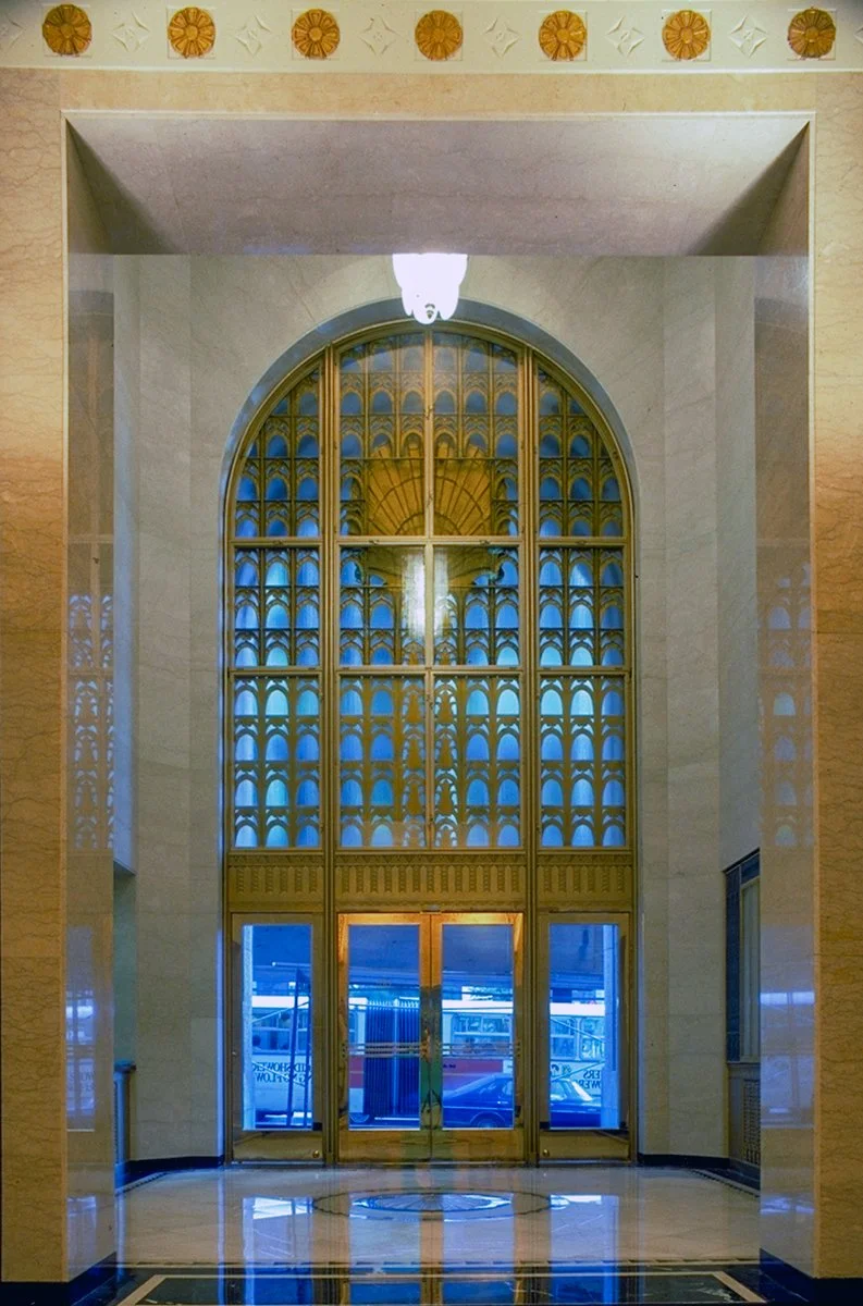 Large golden door with ornate lattice windows, arched top, in a building lobby with marble floors and decorative wall accents.