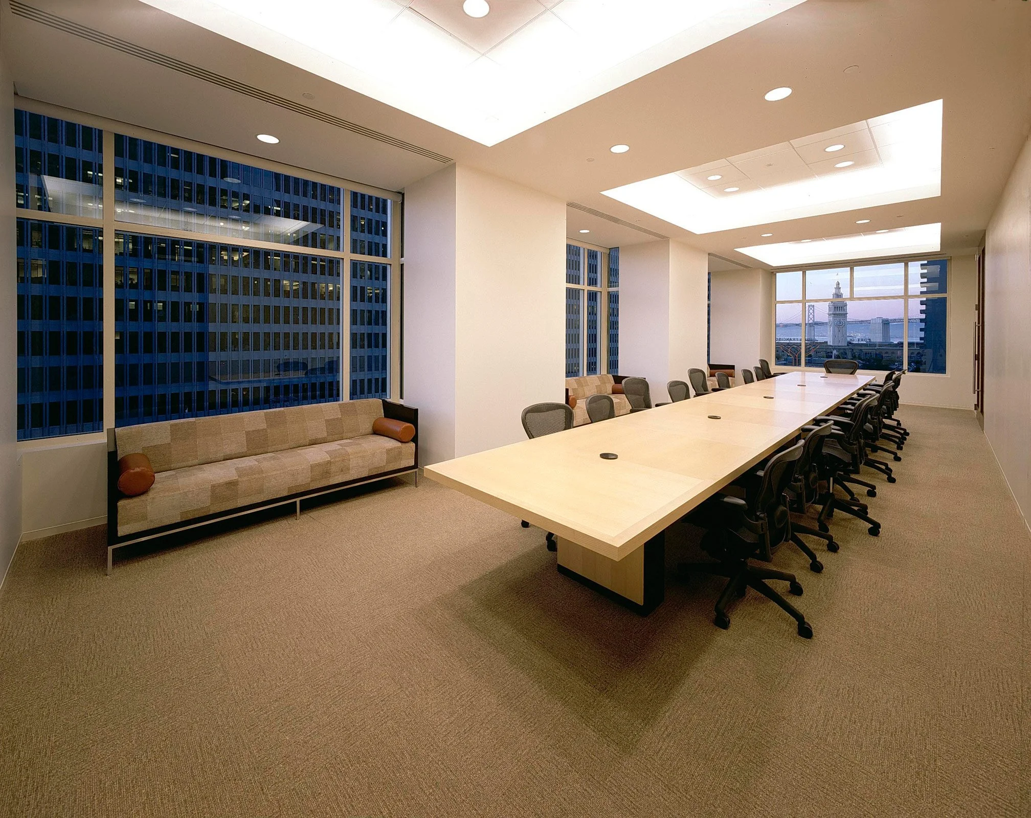 A modern conference room with a long wooden table surrounded by black office chairs, a beige sofa with pillows along the windowed wall, and large windows showing a cityscape with a tower in the distance, illuminated ceiling lights, and a neutral carp