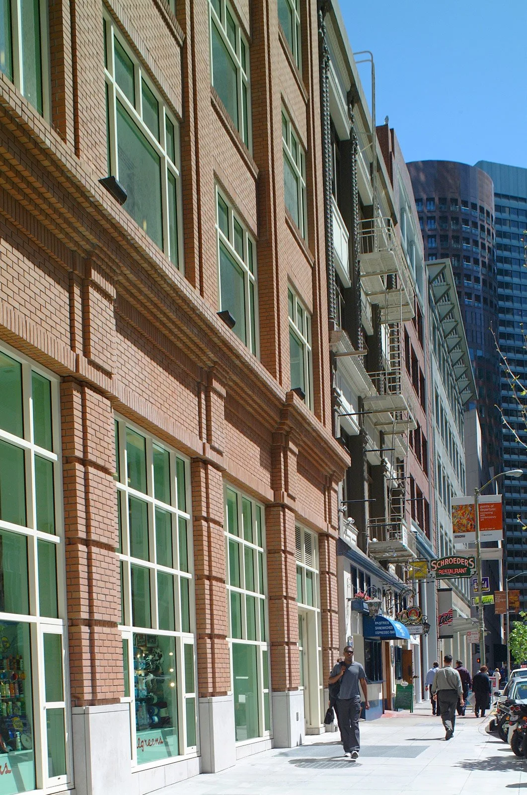 City street scene with pedestrians walking past brick and glass buildings, signs for restaurants, and parked cars.