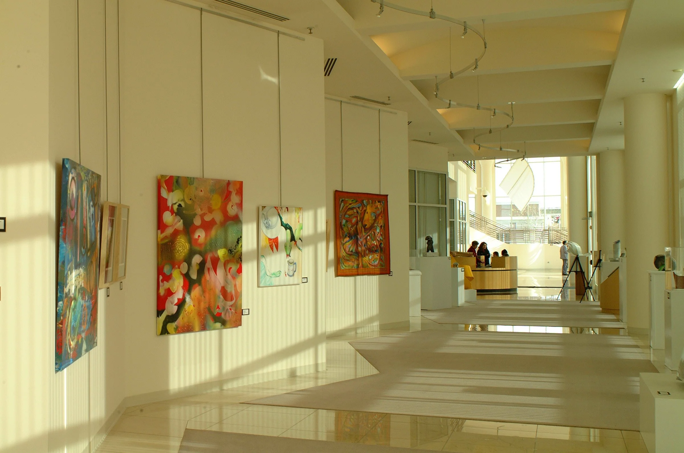 Art gallery interior with colorful paintings hanging on a white wall, sunlight casting shadows, and visitors sitting in the background.