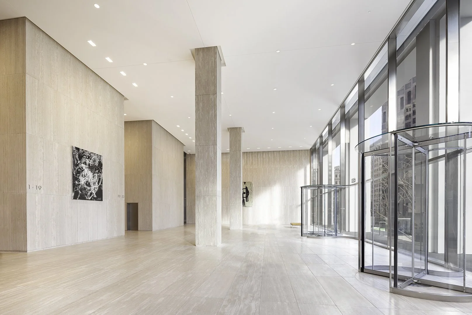 Modern art gallery with beige wooden walls, light hardwood floors, tall glass windows, and a revolving glass door. Art paintings are displayed on the walls.