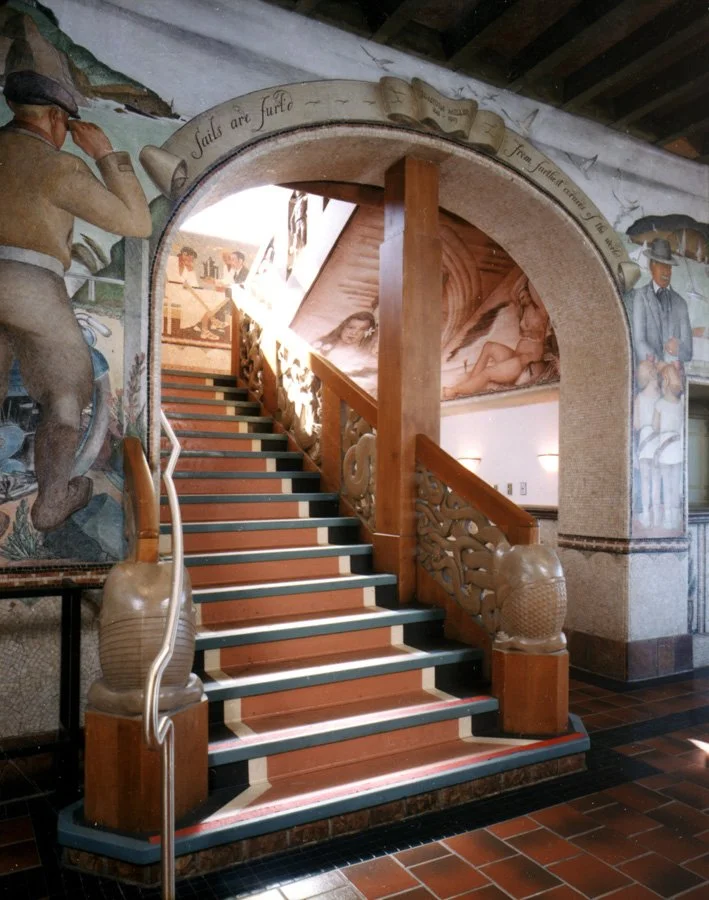 Indoor staircase with wooden handrails and decorative posts, leading up through an archway adorned with murals depicting historical scenes and figures.
