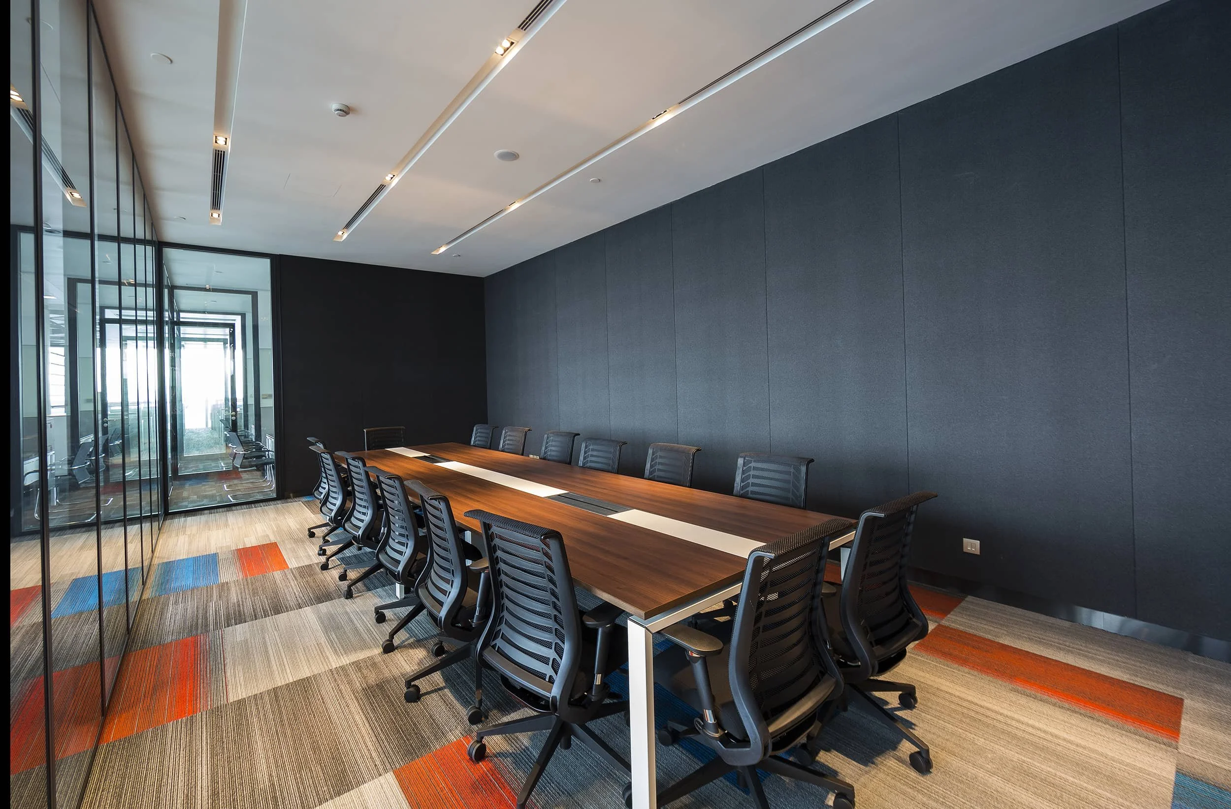 Empty modern conference room with a long wooden table, black chairs, glass walls, and colorful carpet.
