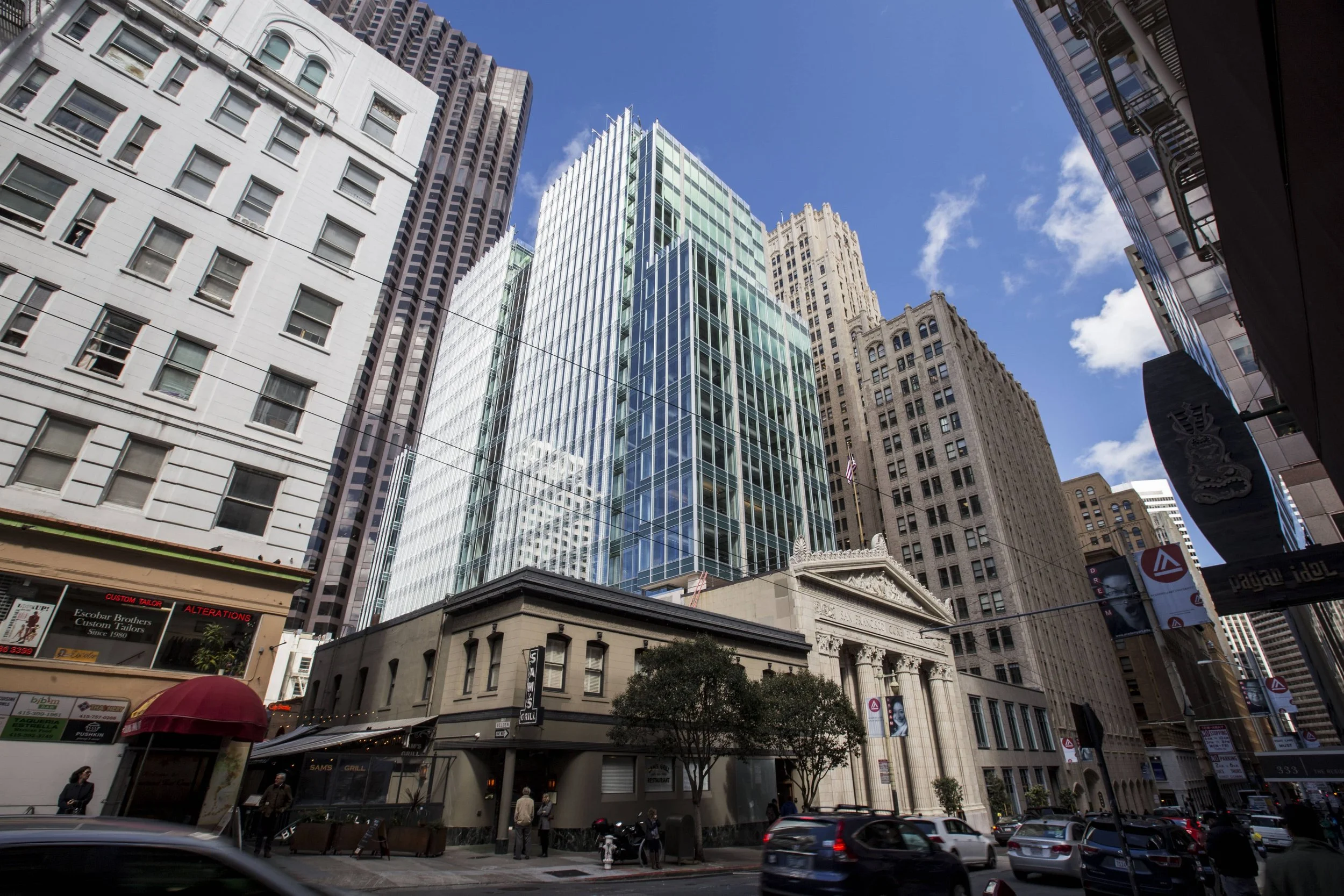 Tall modern glass skyscrapers in a city street scene with cars and pedestrians, blue sky with some clouds.
