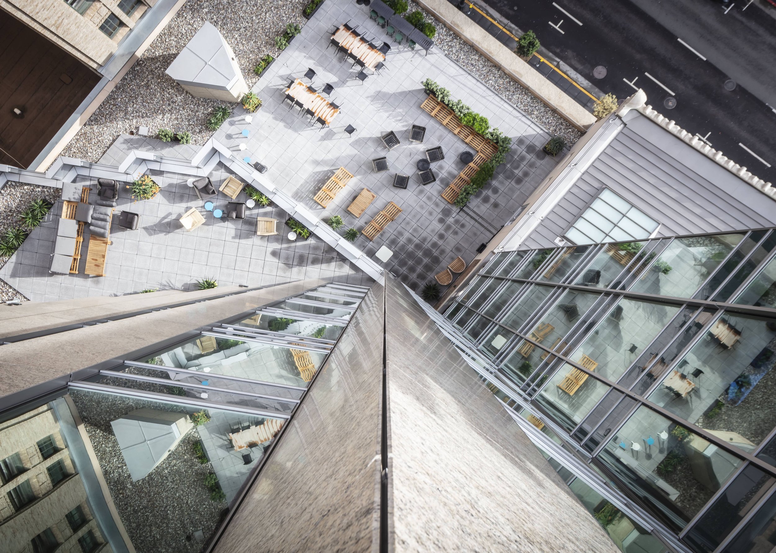 A high-angle view of a rooftop terrace with outdoor seating, tables, chairs, benches, and plants, surrounded by glass building walls and overlooking the street.