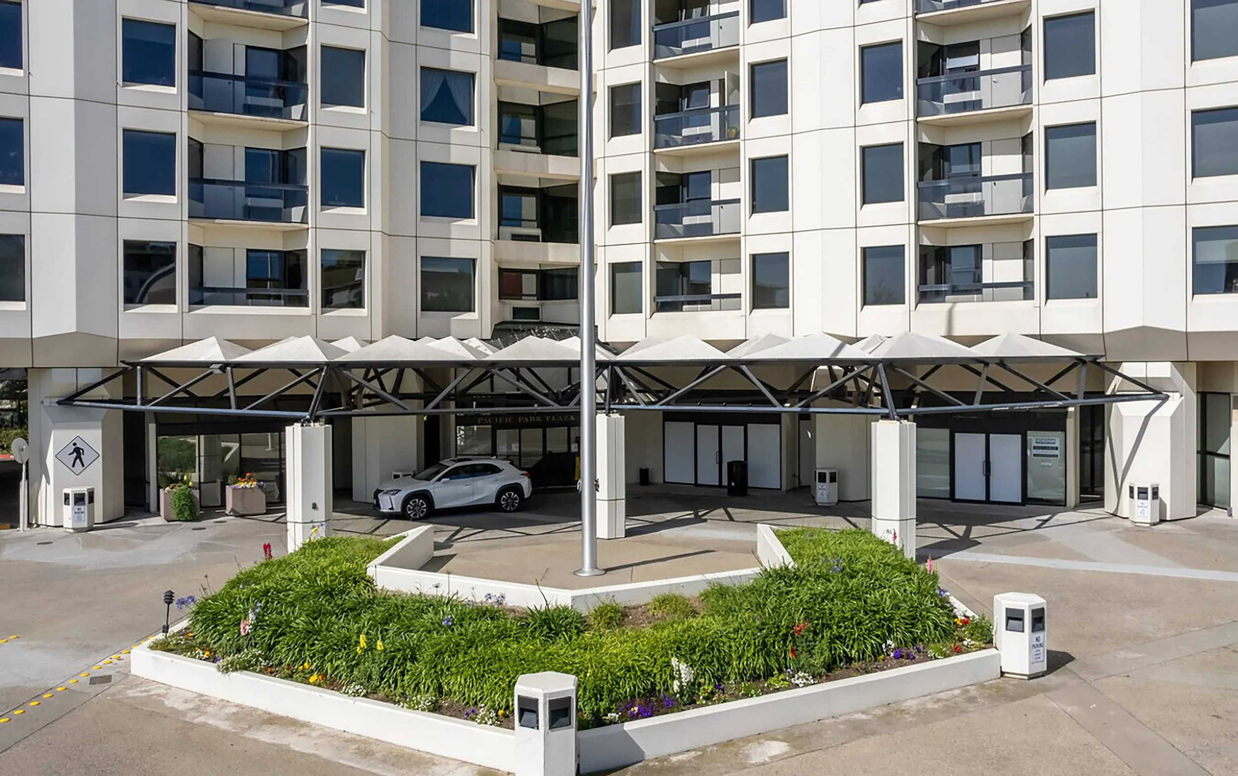 View of a modern apartment building entrance with a white car parked under a canopy, landscaped flower bed in front, and a flagpole in the center.