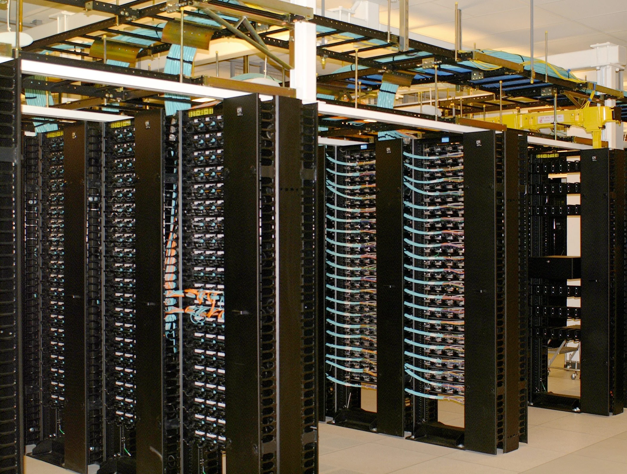 A large server room with rows of black server racks filled with computer equipment. The room has overhead lighting and ceiling-mounted cabling management.