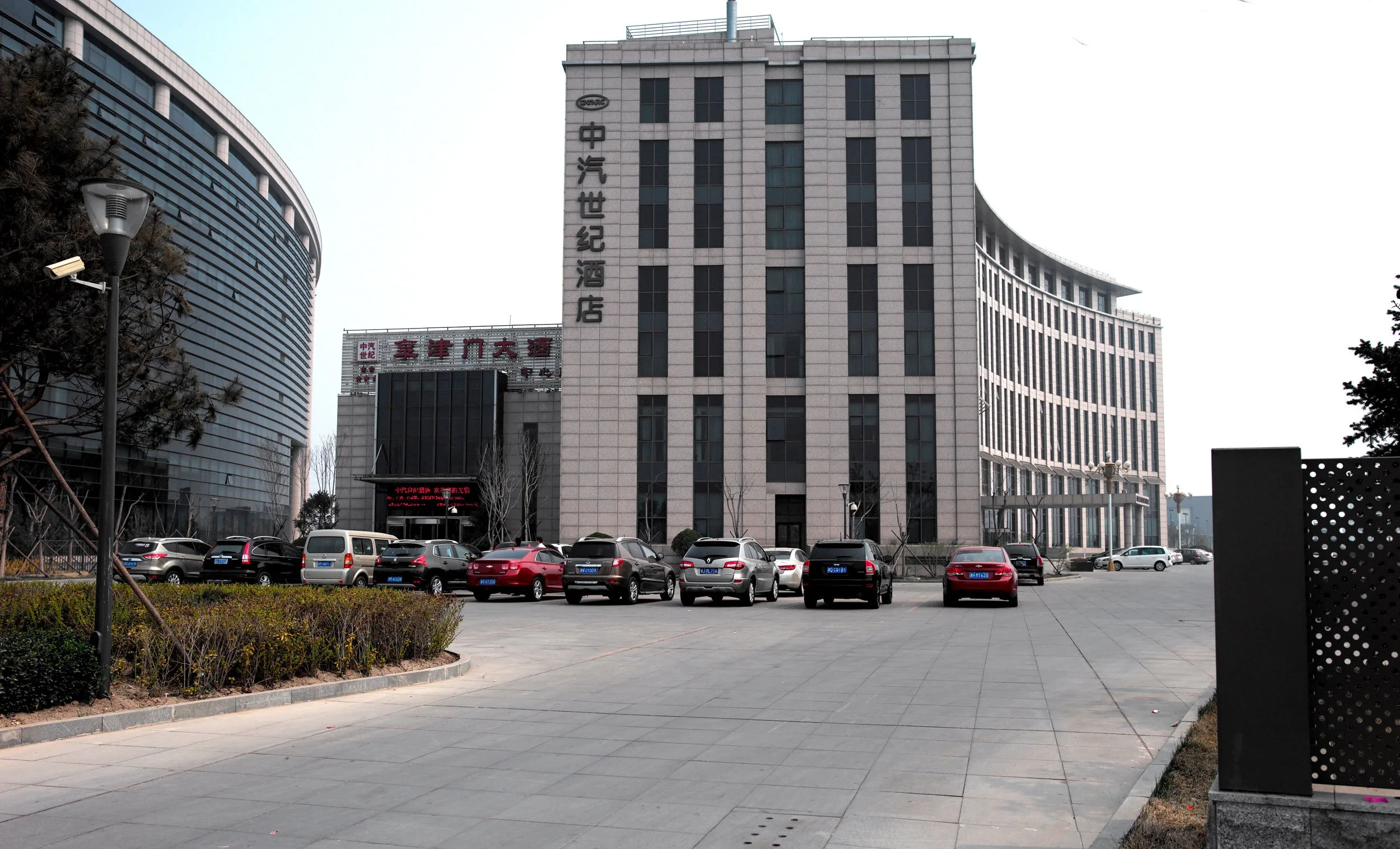 View of a parking lot in front of modern commercial buildings, some with Chinese signage, under a cloudy sky.