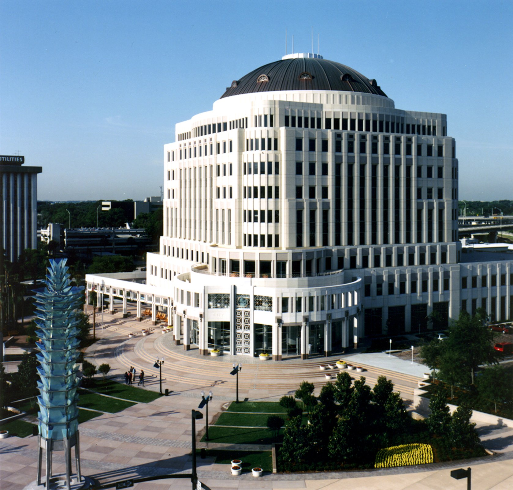 A large, white multi-story building with a rounded roof and numerous windows, situated in an urban area with a plaza, trees, and sculptures in the foreground.