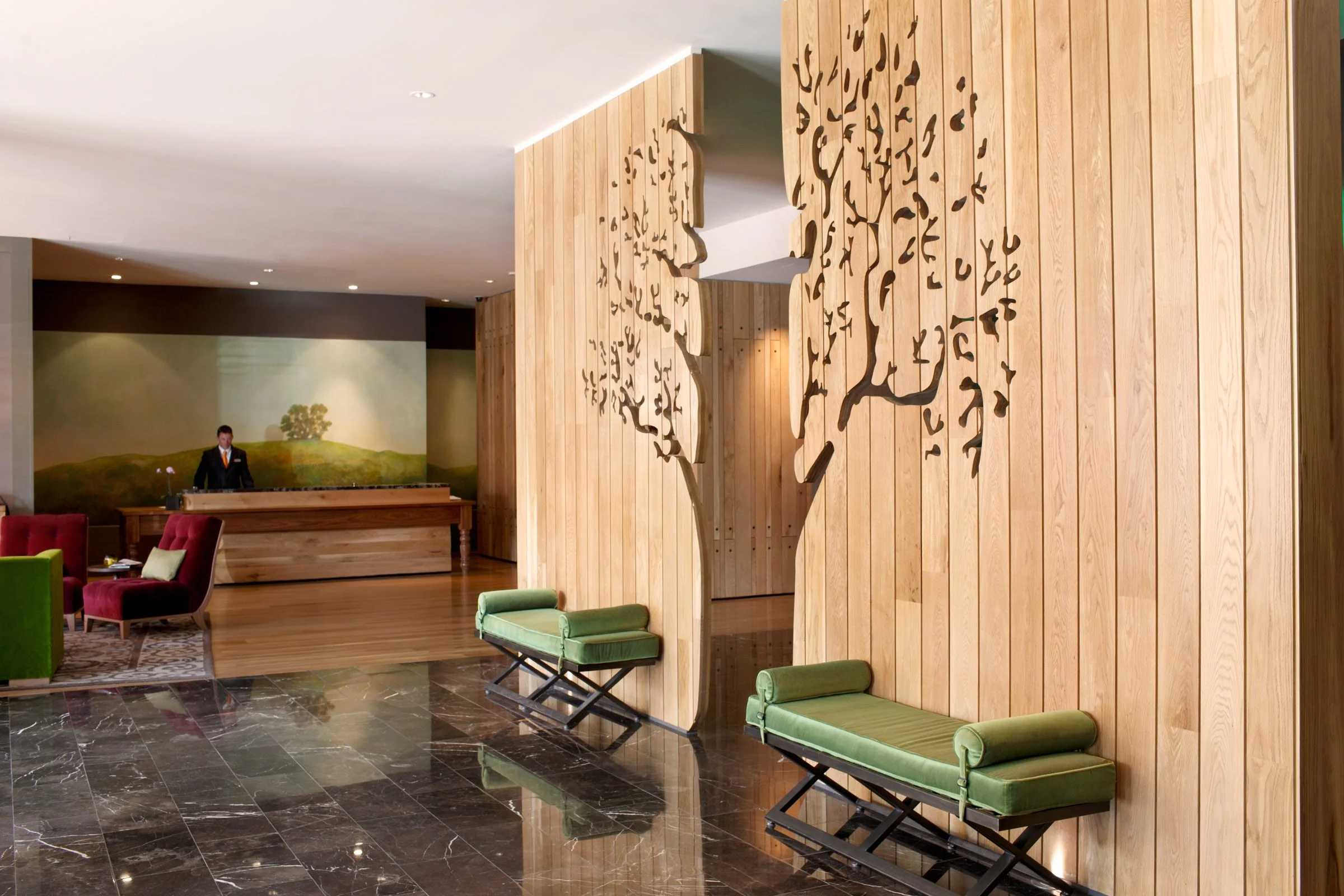 Elegant hotel lobby with a wooden wall featuring tree branch cutouts, green benches, a wooden reception desk, and a decorative landscape painting in the background.