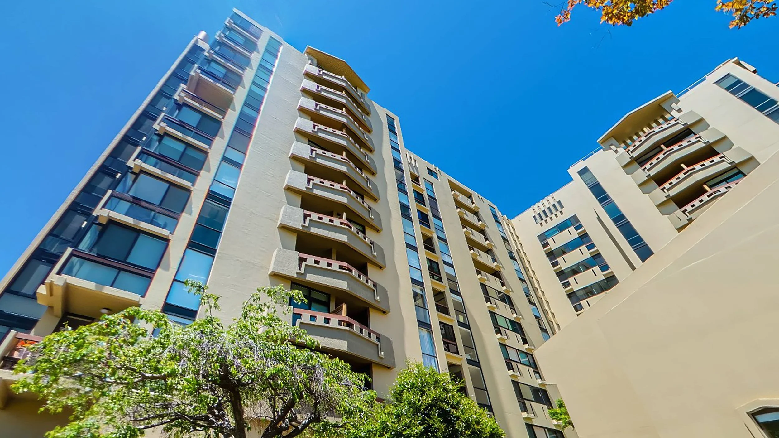 Looking up at a tall modern apartment building with multiple balconies and large windows, set against a clear blue sky with some tree branches in the foreground.
