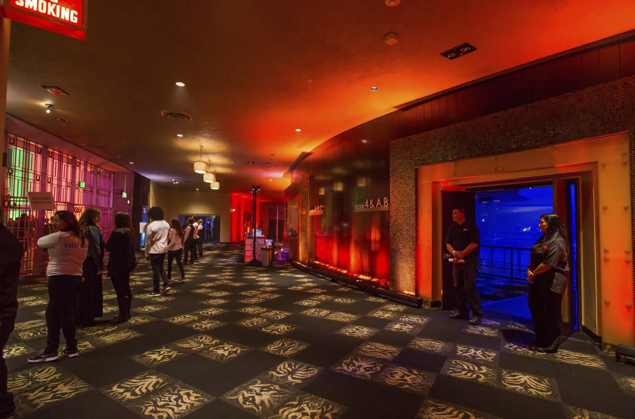 People standing in line inside a dimly lit, colorful event venue with patterned carpet, illuminated red and blue walls, and a bar in the background.