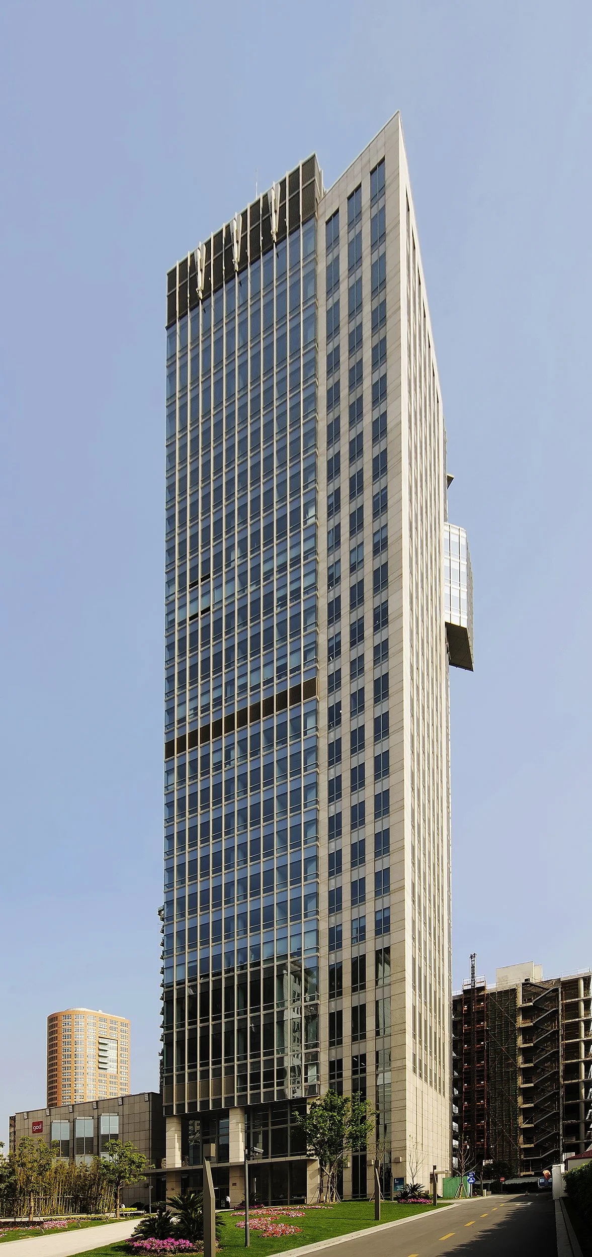 A tall, modern skyscraper with glass windows and vertical metal beams, situated in an urban area with green landscaping at its base.