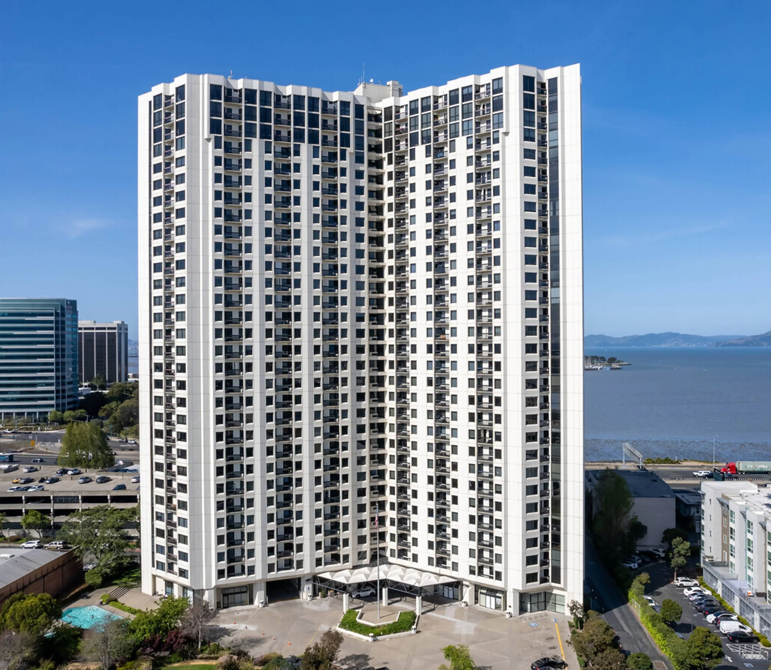 Tall white apartment building with numerous balconies, overlooking a body of water with a scenic view of the coastline and other buildings.