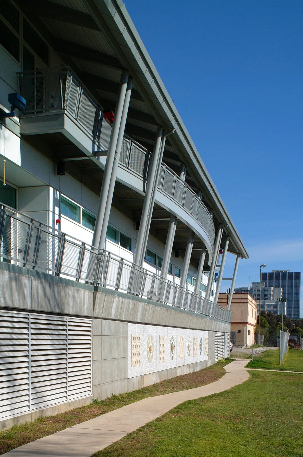 Side view of a modern, multi-story building with metal railings, large windows, and a curved design, surrounded by a grassy area and a walking path, under a clear blue sky.