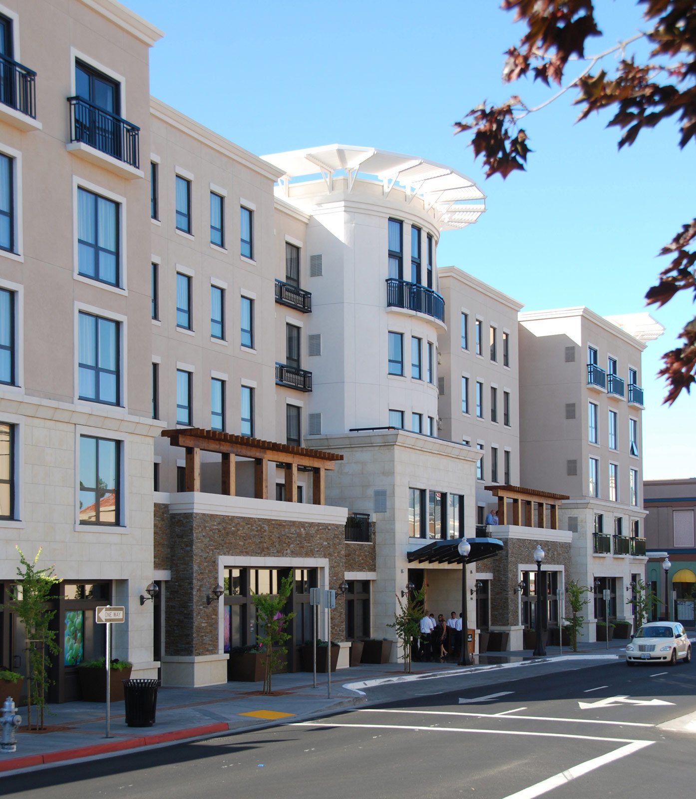 Modern multi-story residential building with beige exterior, black balconies, and large windows, located on a city street with trees, street signs, and people standing outside under a clear blue sky.