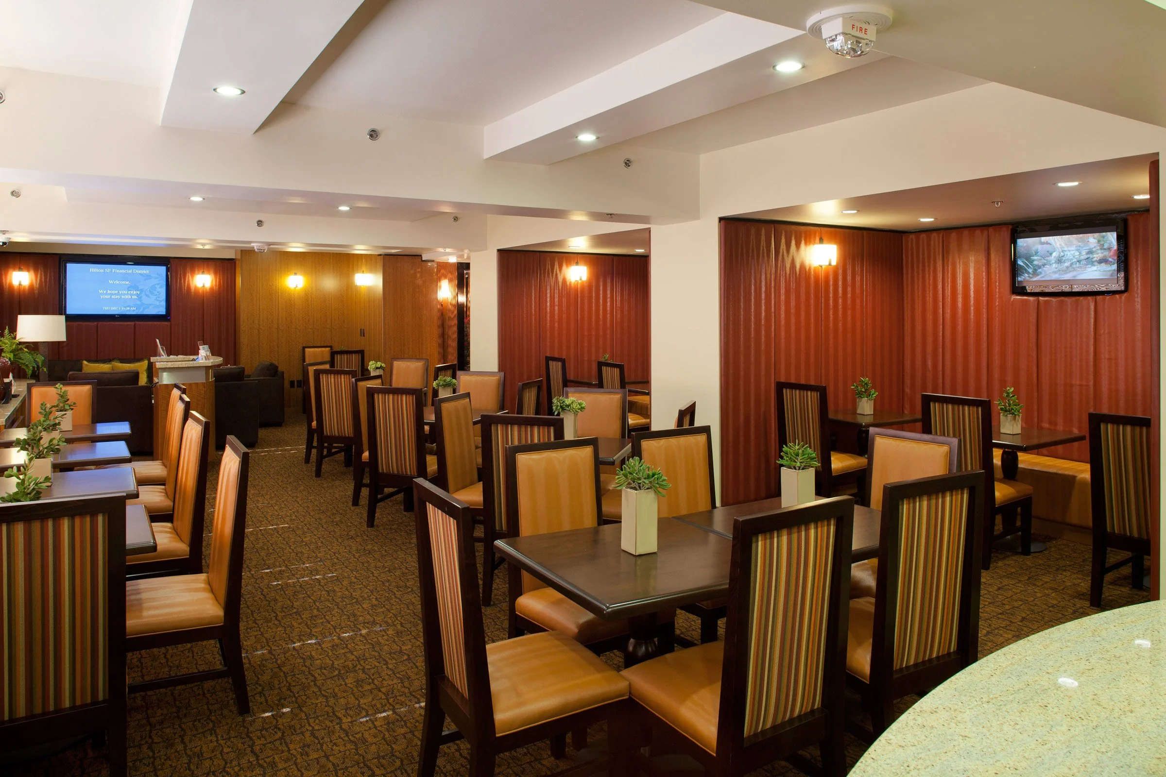A well-lit restaurant interior with multiple tables and chairs, small potted plants on each table, and wall-mounted flat-screen TVs.