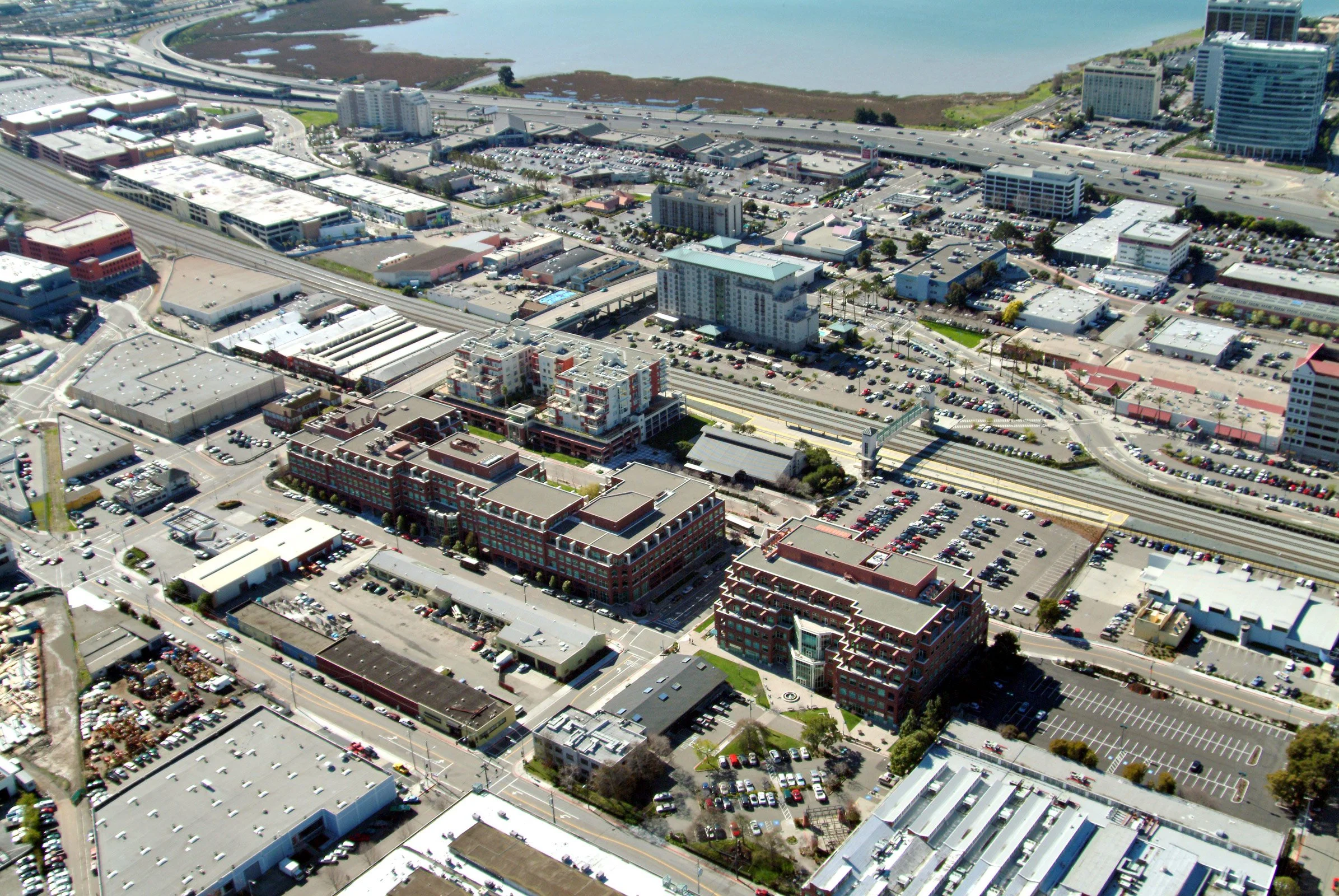 Aerial view of a cityscape near a water body, showing commercial buildings, parking lots, roads, and railways.