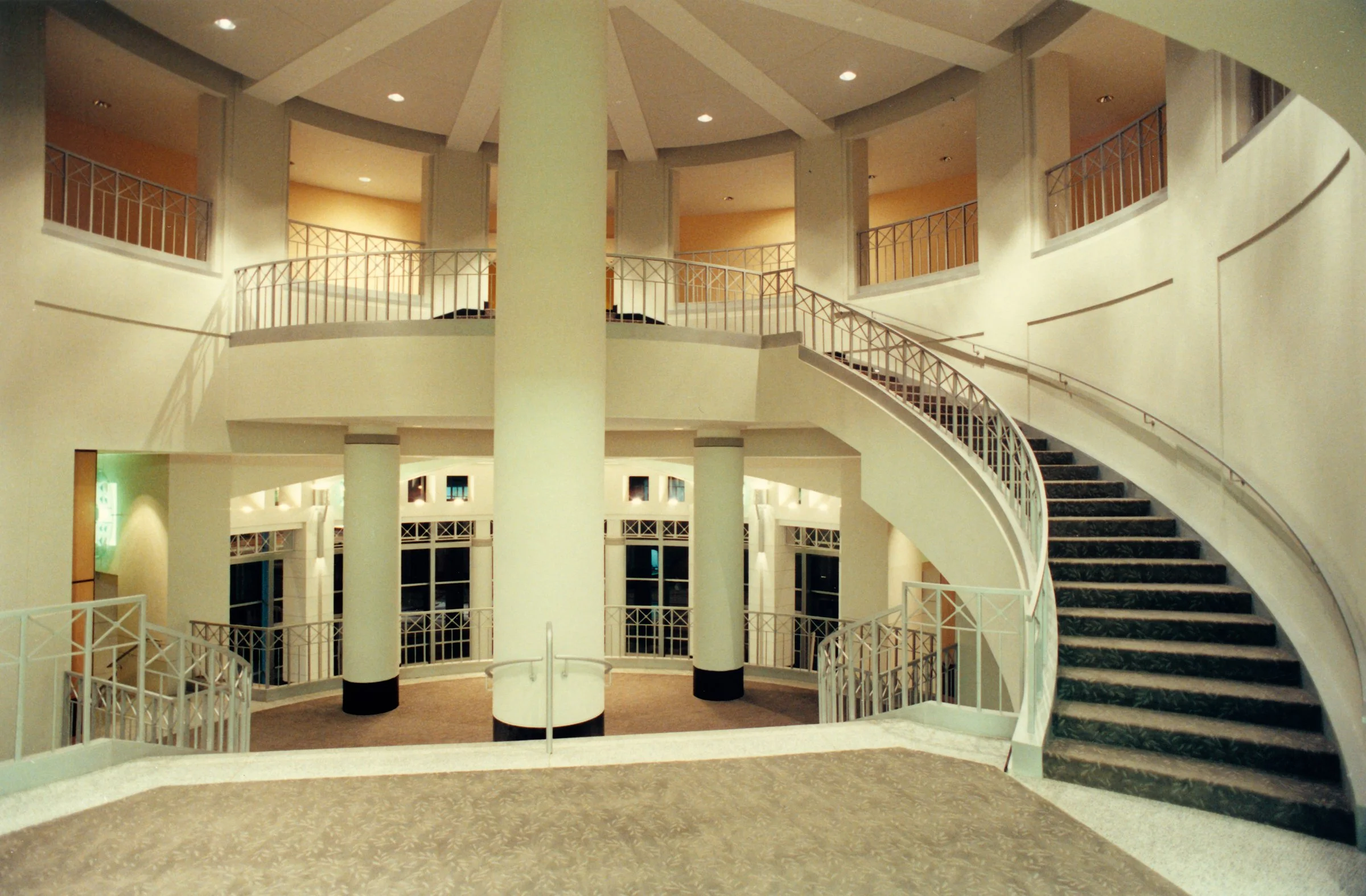 Interior view of a spacious, multi-level hotel lobby with a curved staircase, white railings, and tall white columns, illuminated with warm lighting.