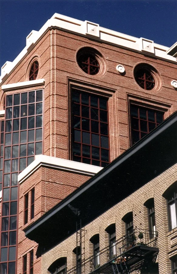 Close-up of a brick building with large windows, decorative round windows with crosses, and fire escapes, against a clear blue sky.