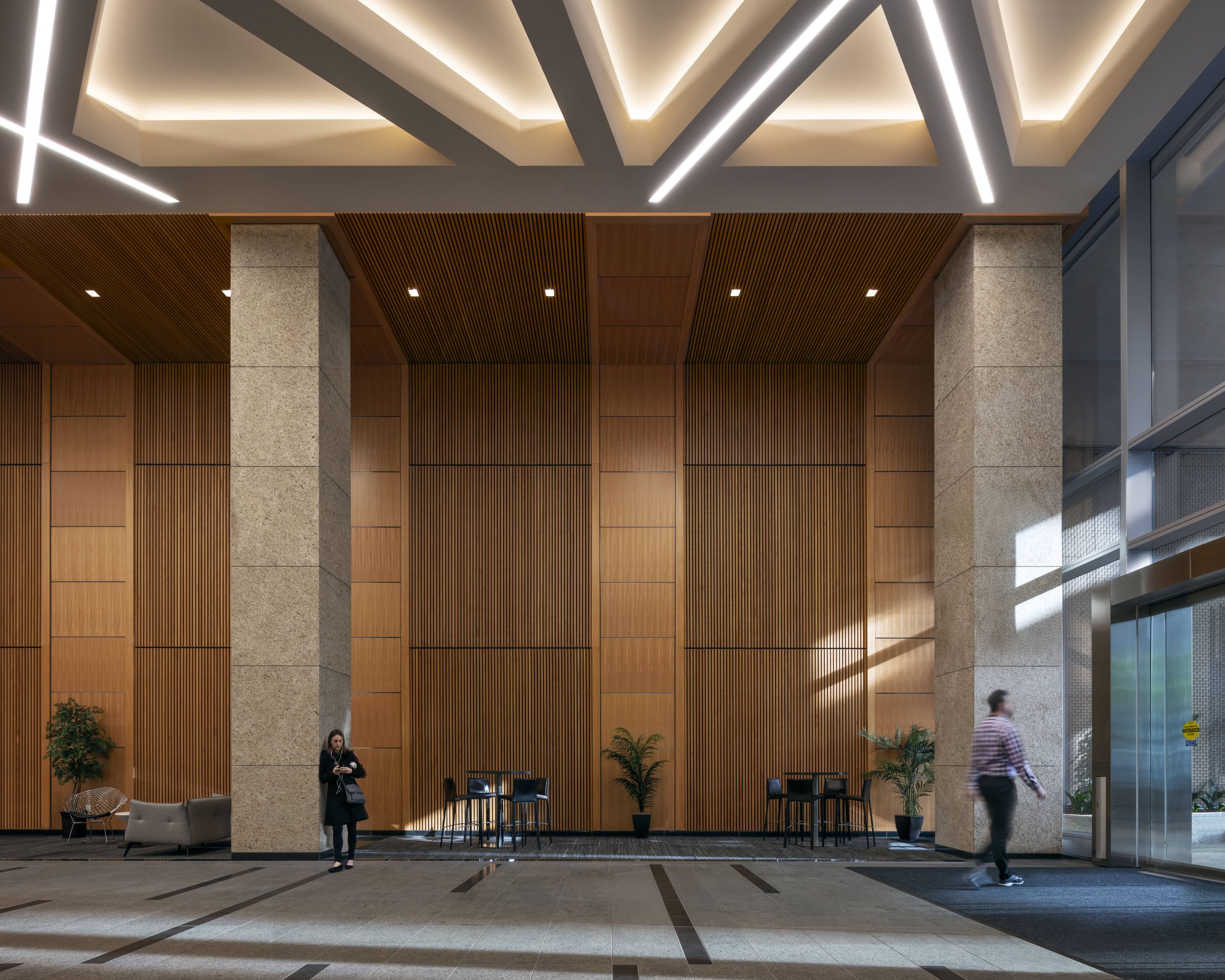 Modern hotel or office lobby with wood-paneled walls, large concrete pillars, potted plants, and seating area, with a woman standing and a man walking near the entrance.