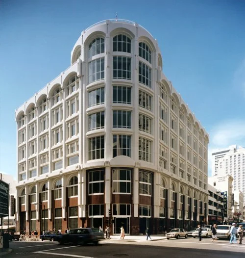 A large, white, multi-story building with rounded corners and numerous windows, situated on a city street with cars and pedestrians.