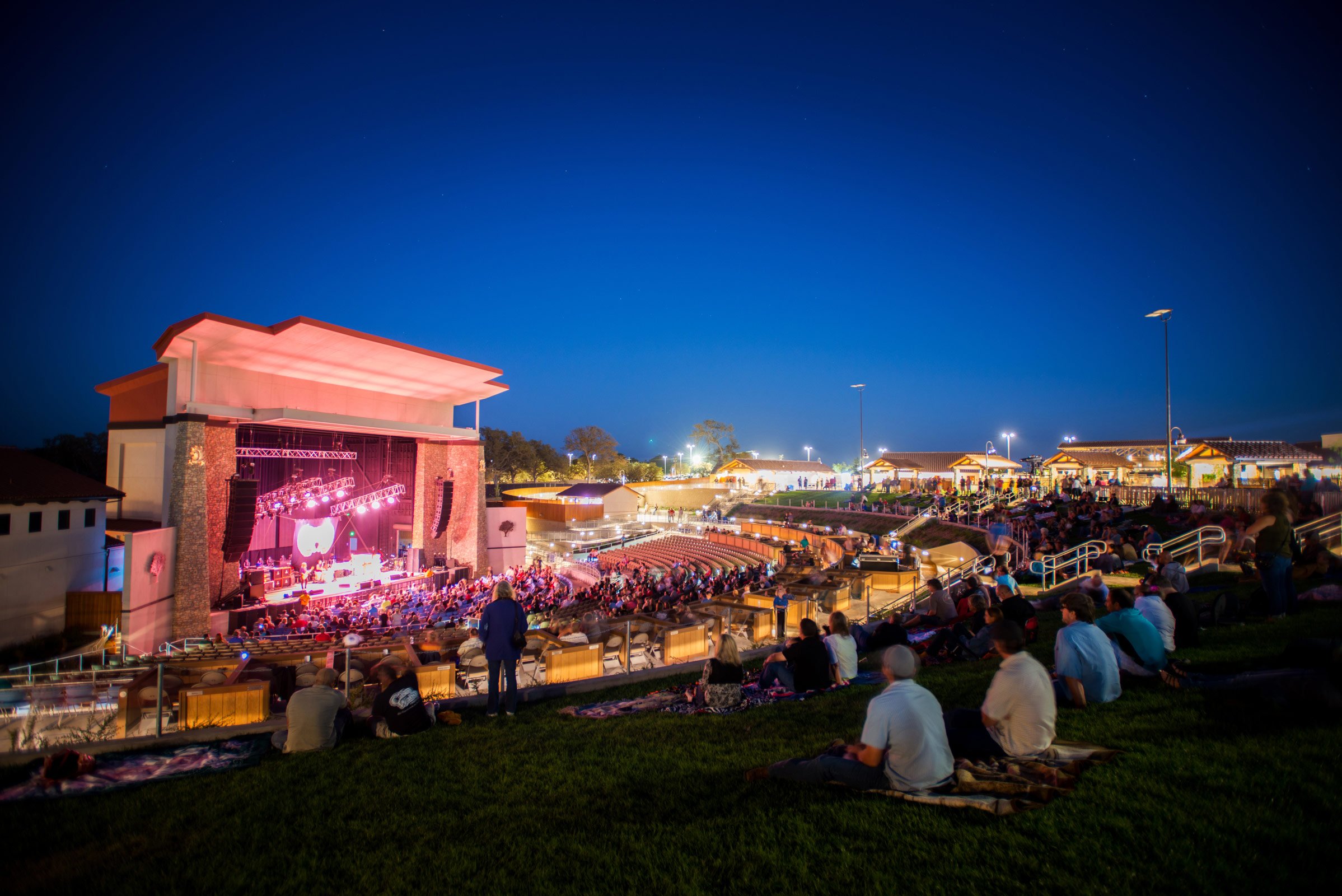 An outdoor amphitheater at dusk with a large crowd watching a concert, illuminated stage, grassy seating area, and surrounding buildings with lights.