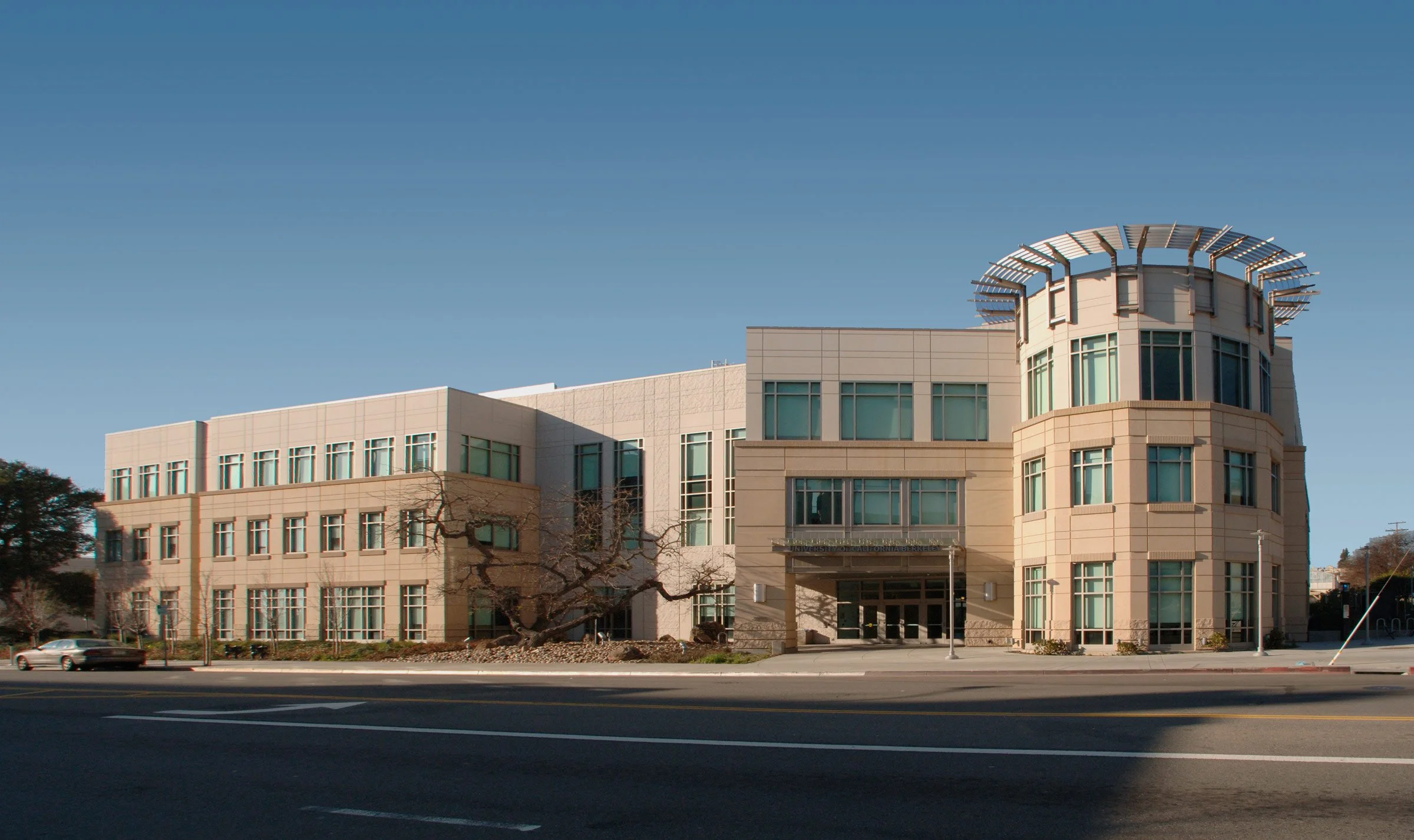 A modern multi-story building with large windows and a unique round tower with a decorative roof, situated along a street with a few cars and trees in front, under a clear blue sky.